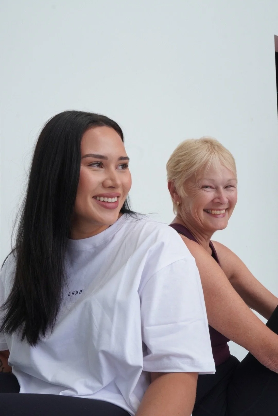 Two women sitting in front of a white wall while practicing Pilates at Refinery Studios, smiling and looking to their left. One woman has long dark hair, and the other has short blonde hair.