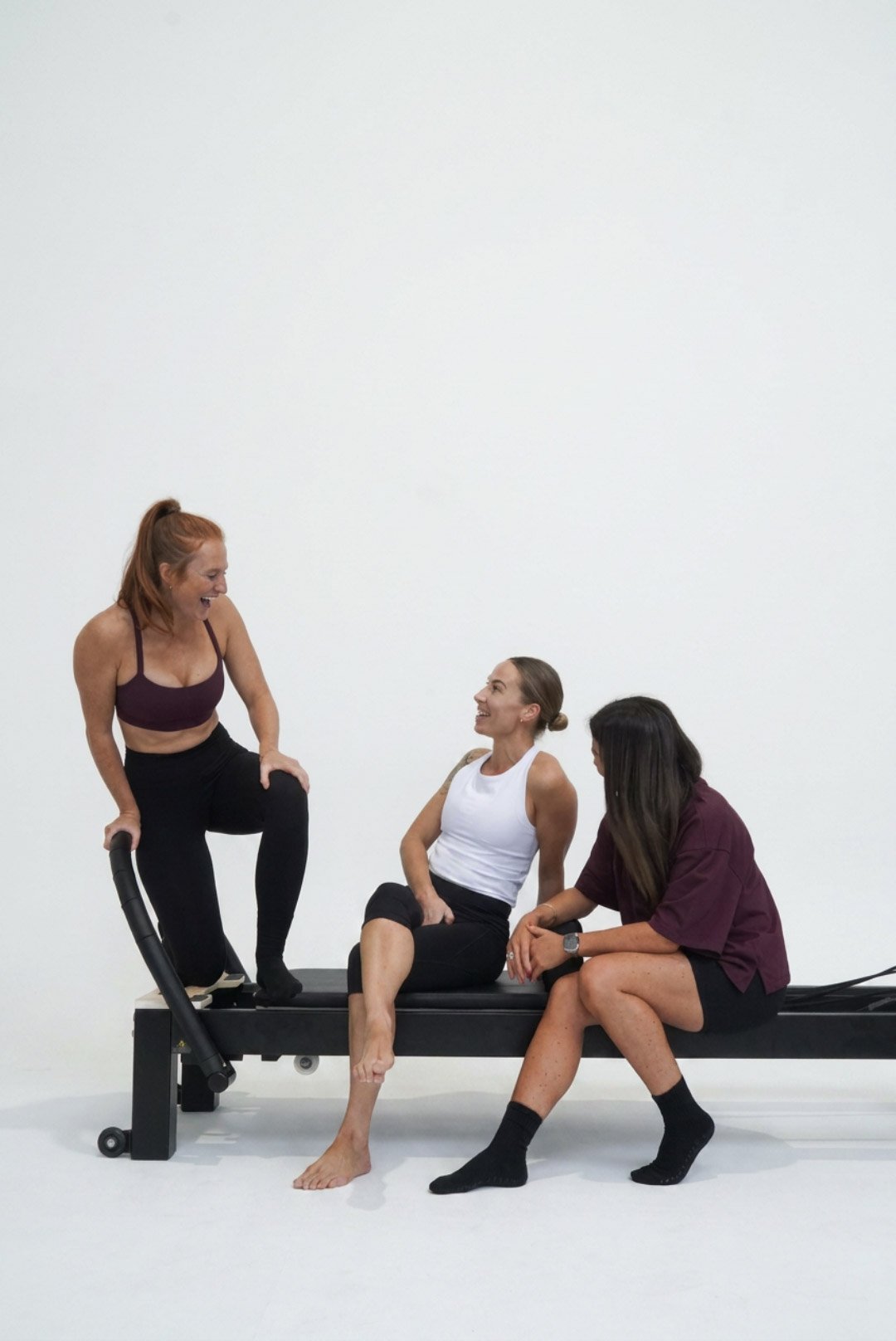 Three women in a Refinery Studios Pilates classes, with one standing and smiling, two sitting on the reformer, all engaging in conversation.