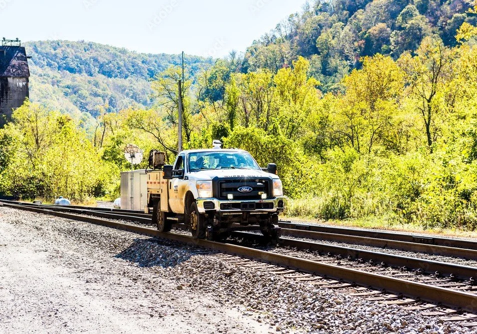 A white Ford utility truck on railroad tracks with green trees and hills in the background.