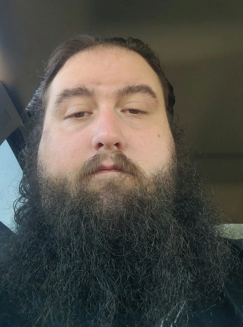 Close-up of a man with long dark hair and a large beard looking into the camera inside a vehicle.