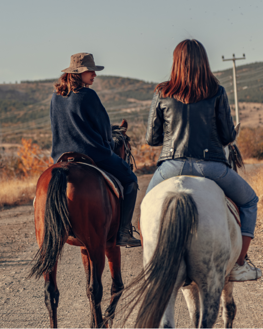 Two women riding horses on a dirt path in a rural landscape with hills and sparse vegetation, during late afternoon or early evening.
