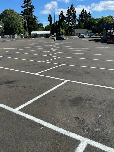 Empty parking lot with white painted parking spaces and a few parked cars in the background, some trees, and a partly cloudy sky.
