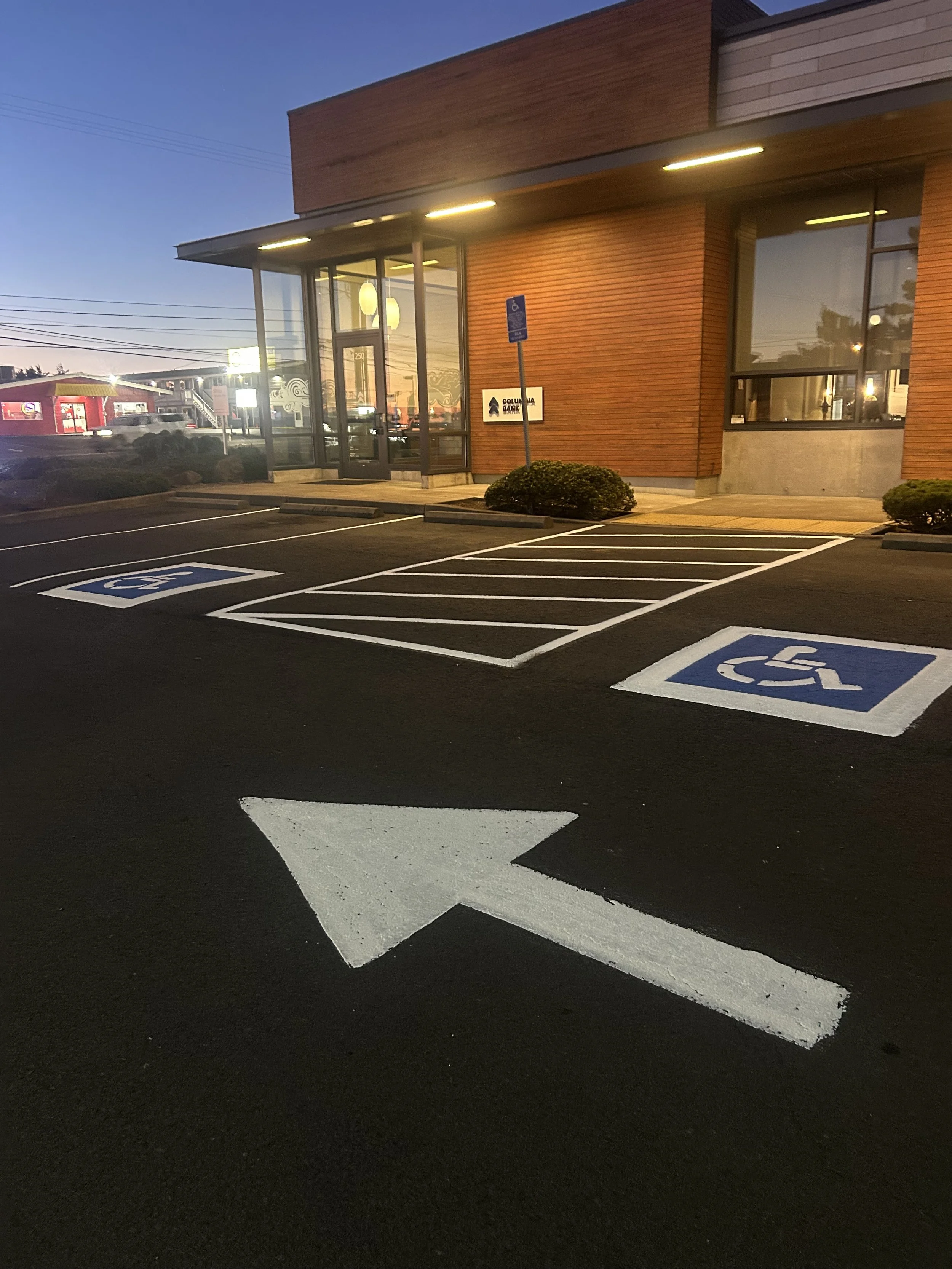 A parking lot at dusk with marked handicap parking spaces and a building with large windows, a sign for Columbia Bank, and a parking meter