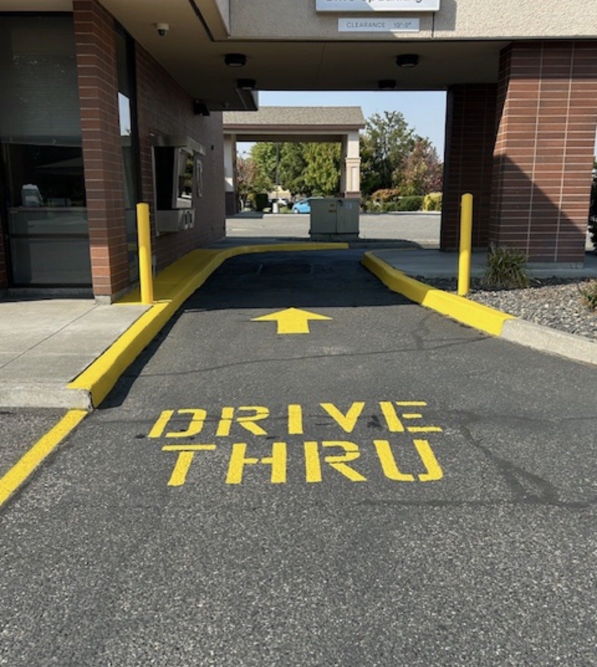 A drive-thru lane with yellow painted text on the pavement that says 'DRIVE THROUGH' and an arrow pointing forward, leading under a building. The lane is bordered by yellow curbs and posts.