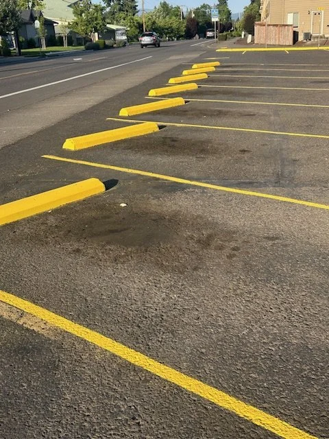 Empty parking lot with yellow parking blocks and lines on asphalt pavement, near a street with cars and buildings in the background.