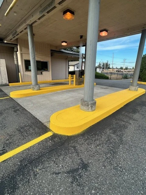 Empty drive-thru lane with yellow painted curb and protective bollard poles under a building with ceiling lights, a window, and a small building in the background.