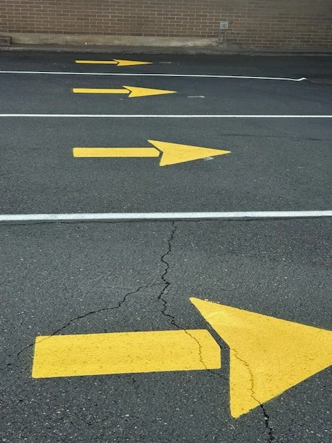 Multiple yellow arrows painted on a parking lot with a crack in the pavement, pointing in the direction of traffic flow.