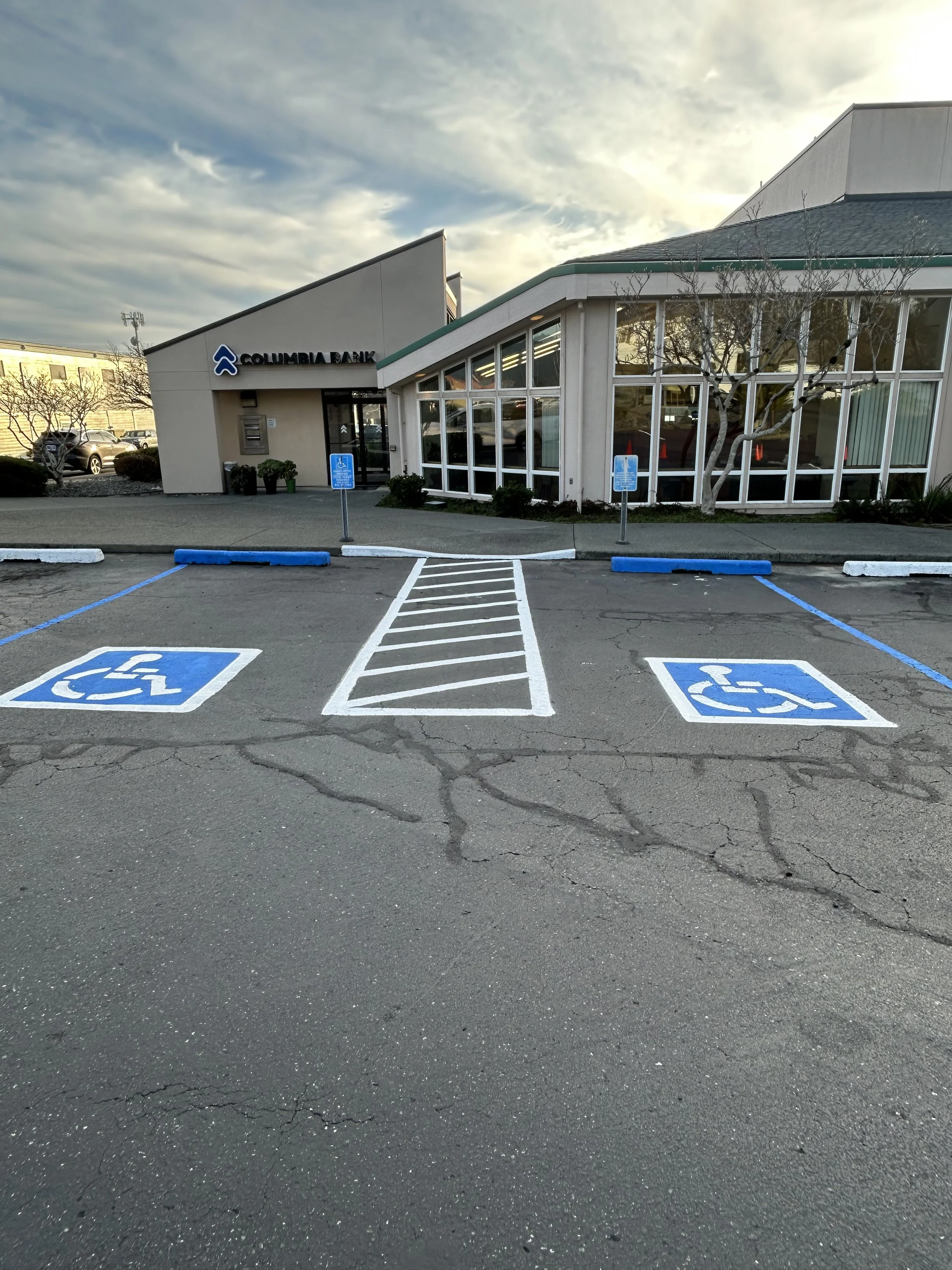 The image shows a parking lot with designated handicapped parking spaces and signs. There are two lease spots in the foreground, marked with blue and white wheelchair symbols. The lot is cracked and slightly worn. In the background, there is a white 