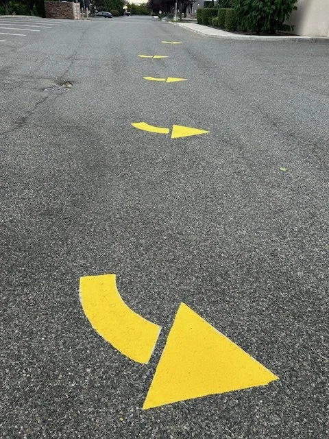 Yellow directional arrows painted on a gray asphalt road, indicating a curve to the left.