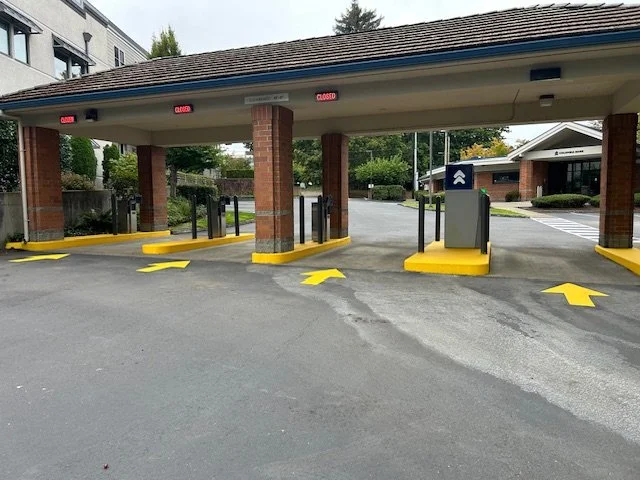 Empty drive-thru with yellow directional arrows on the pavement, security barriers, and closed signs on the overhead structure at a restaurant or retail establishment.