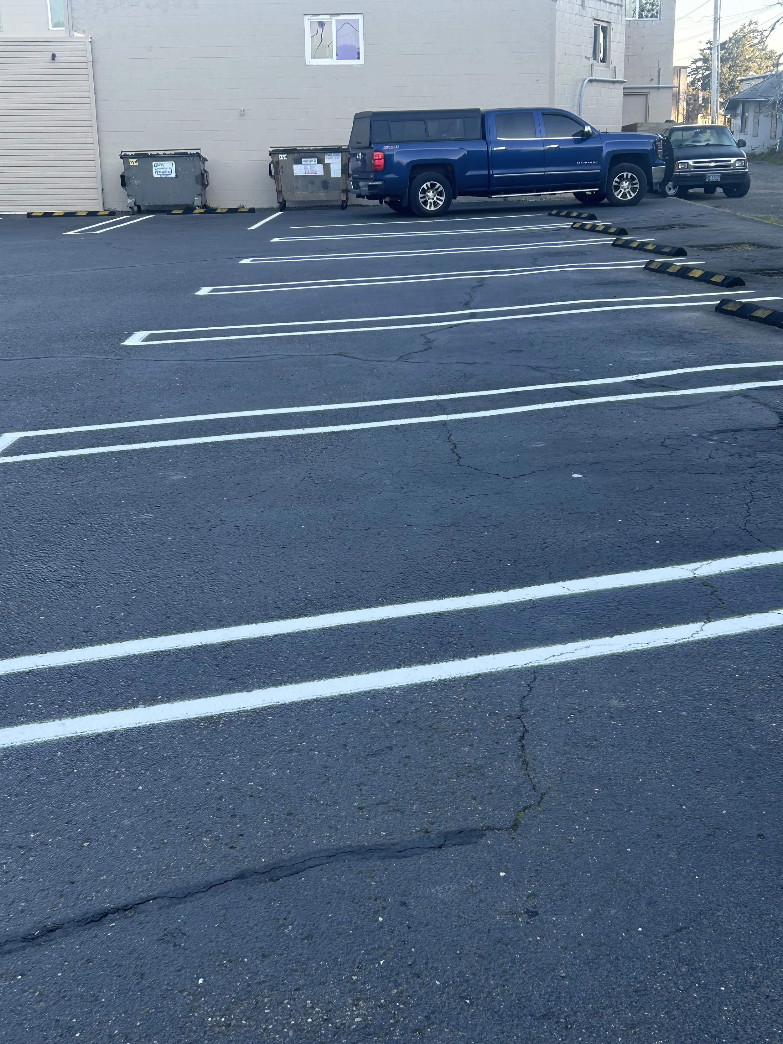 Parking lot with multiple empty parking spaces outlined with white lines. Two dumpsters are against a gray building wall, and a blue pickup truck and a smaller black car are parked along the wall. There are small cracks in the asphalt surface.