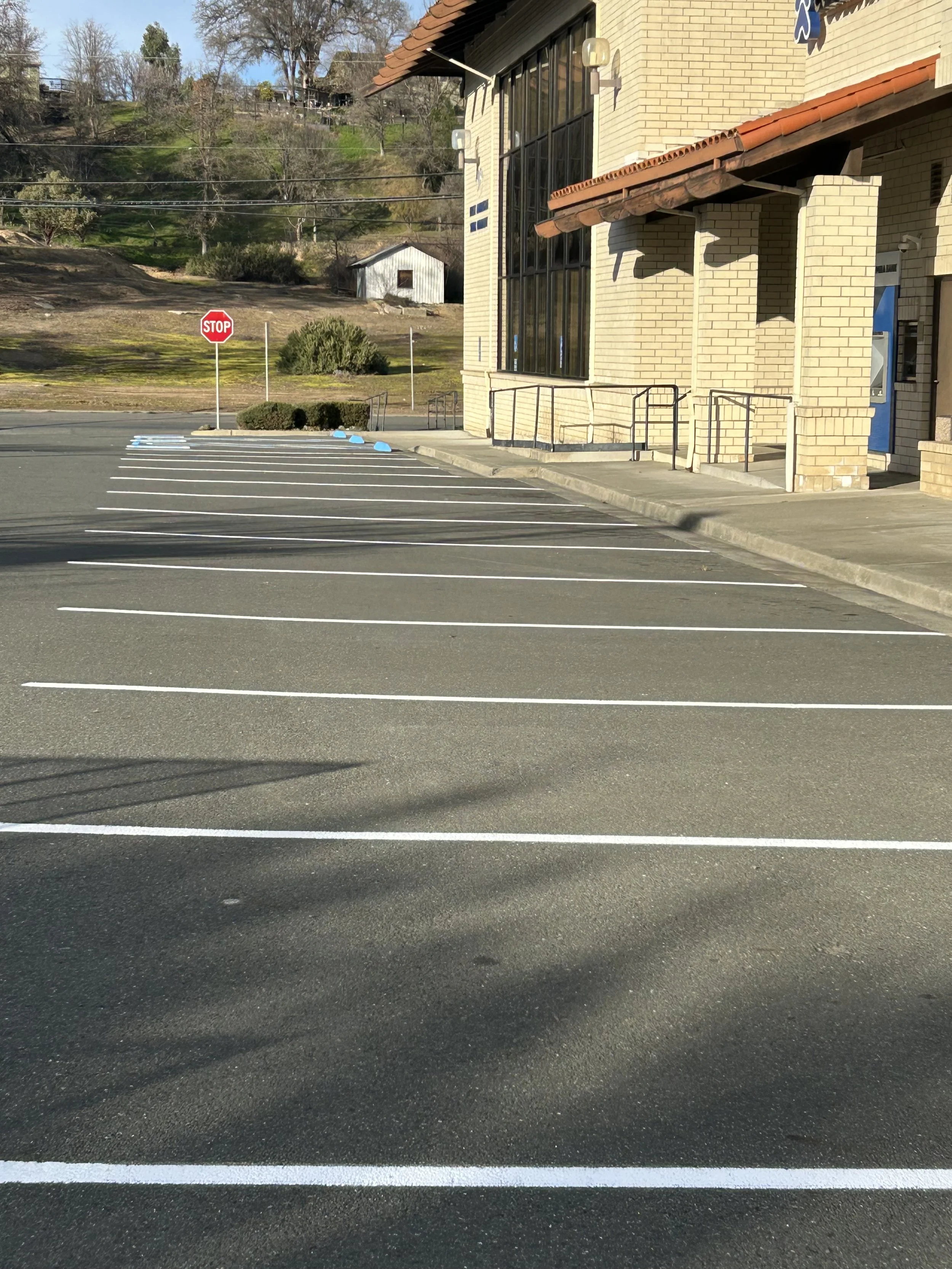 Empty parking lot with white marking lines, a curb, and a corner of a brick building with a blue door. A stop sign is visible in the background against a hillside with trees and a small shed.