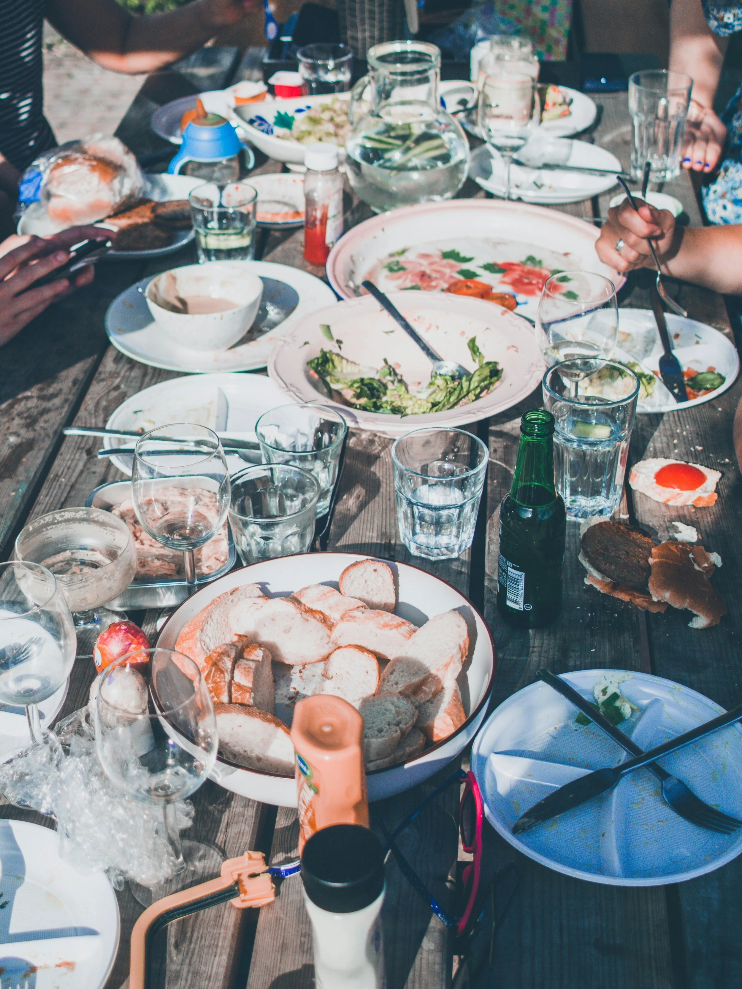 A cluttered outdoor dining table with various partially eaten foods, glasses, and dishes. There are slices of bread, a bowl of salad, a jug of water, and bottles of condiments. Some hands are visible, one holding a fork.