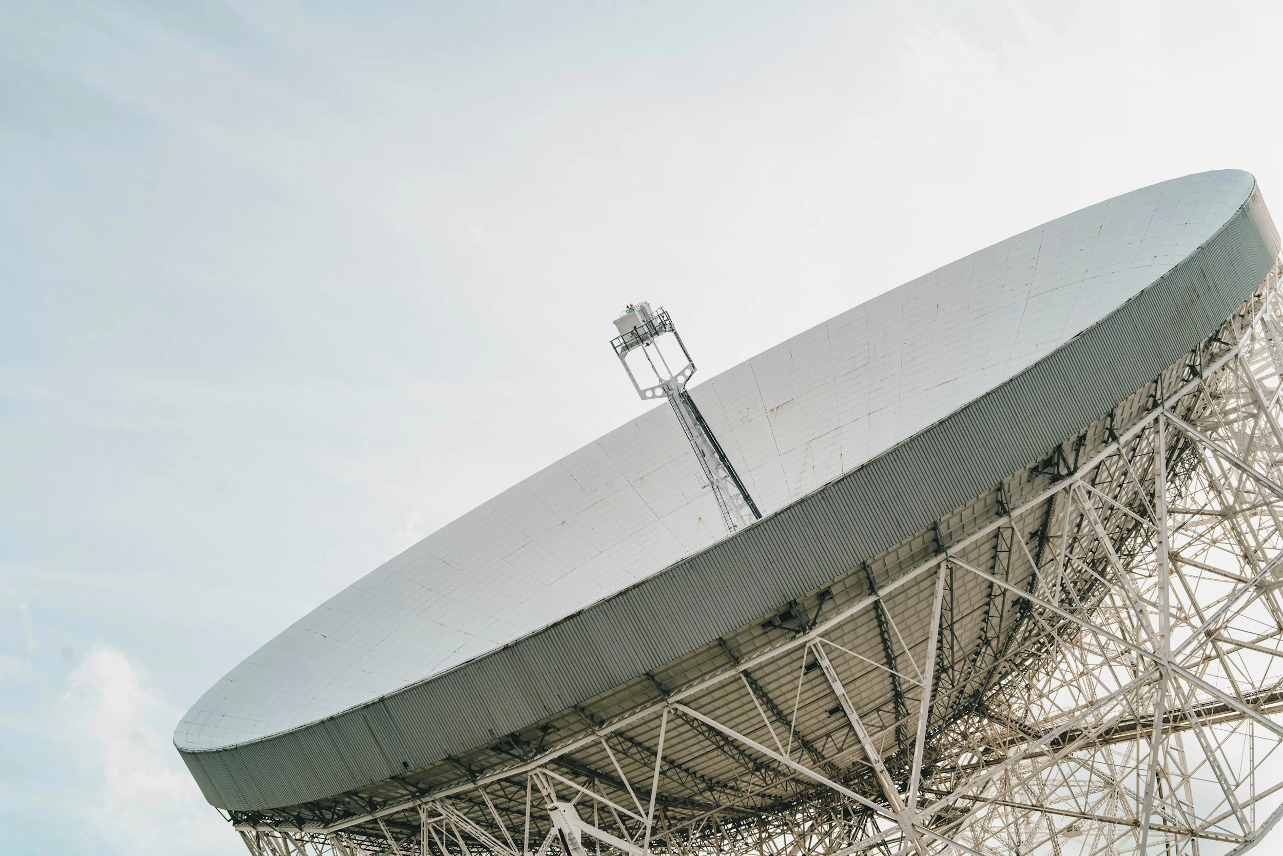 A large satellite dish antenna with a white surface, supported by a metal framework, set against a partly cloudy sky.