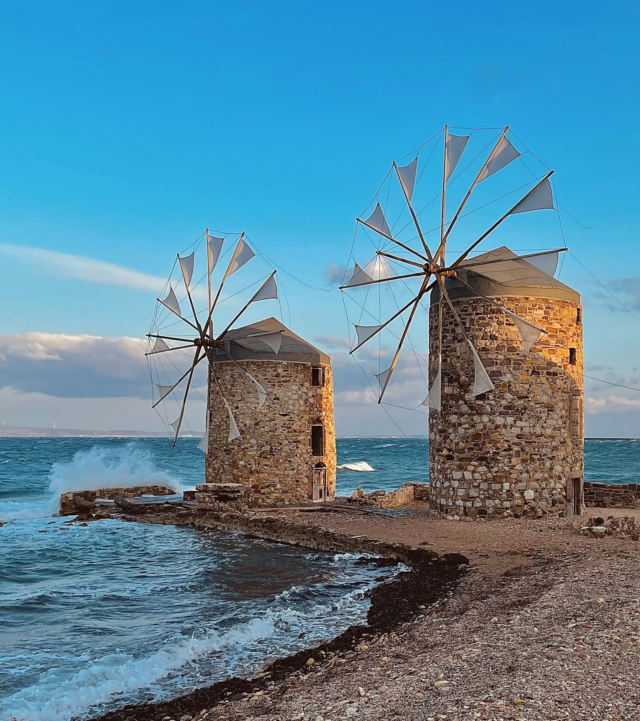 Two old stone windmills with incomplete sail structures on a rocky coast with waves crashing against the rocks, under a blue sky with some clouds.