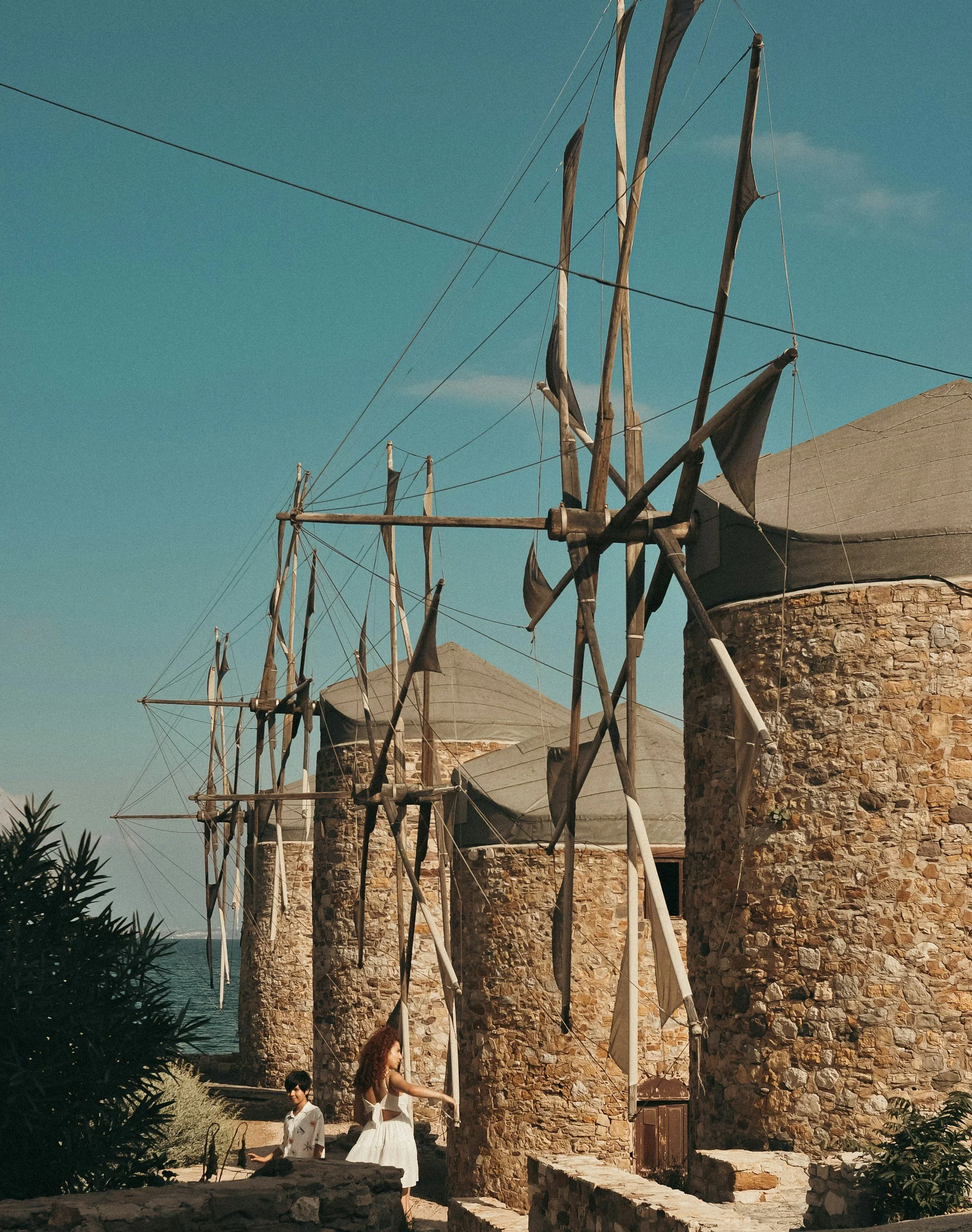 Ancient stone windmills near the coast with black sails, likely in a Mediterranean location, with a woman and a boy near the windmills under a clear blue sky.