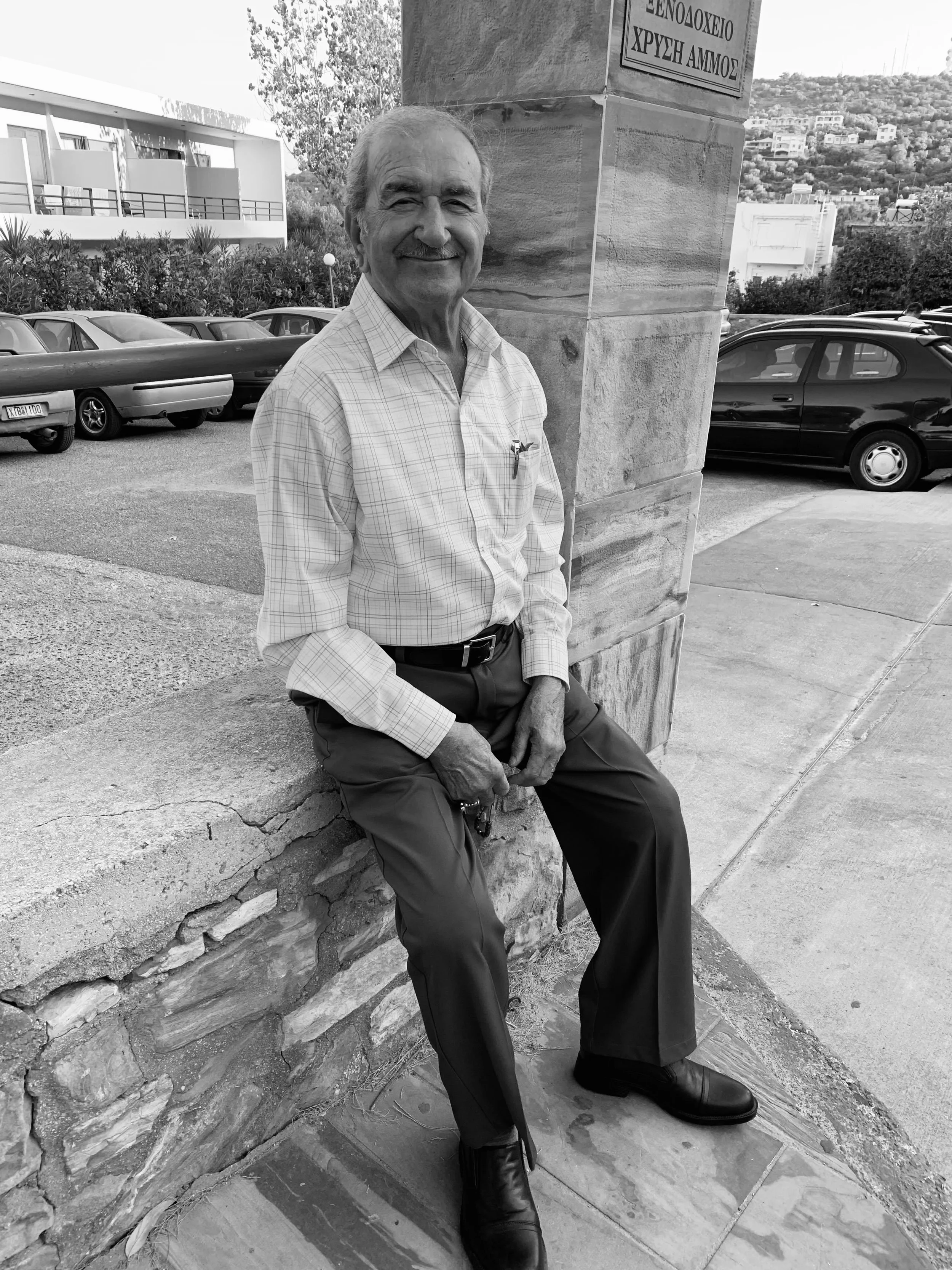 An elderly man with a smile sitting on a stone ledge outside, wearing a plaid shirt, dark pants, and shoes, with parked cars and buildings in the background.
