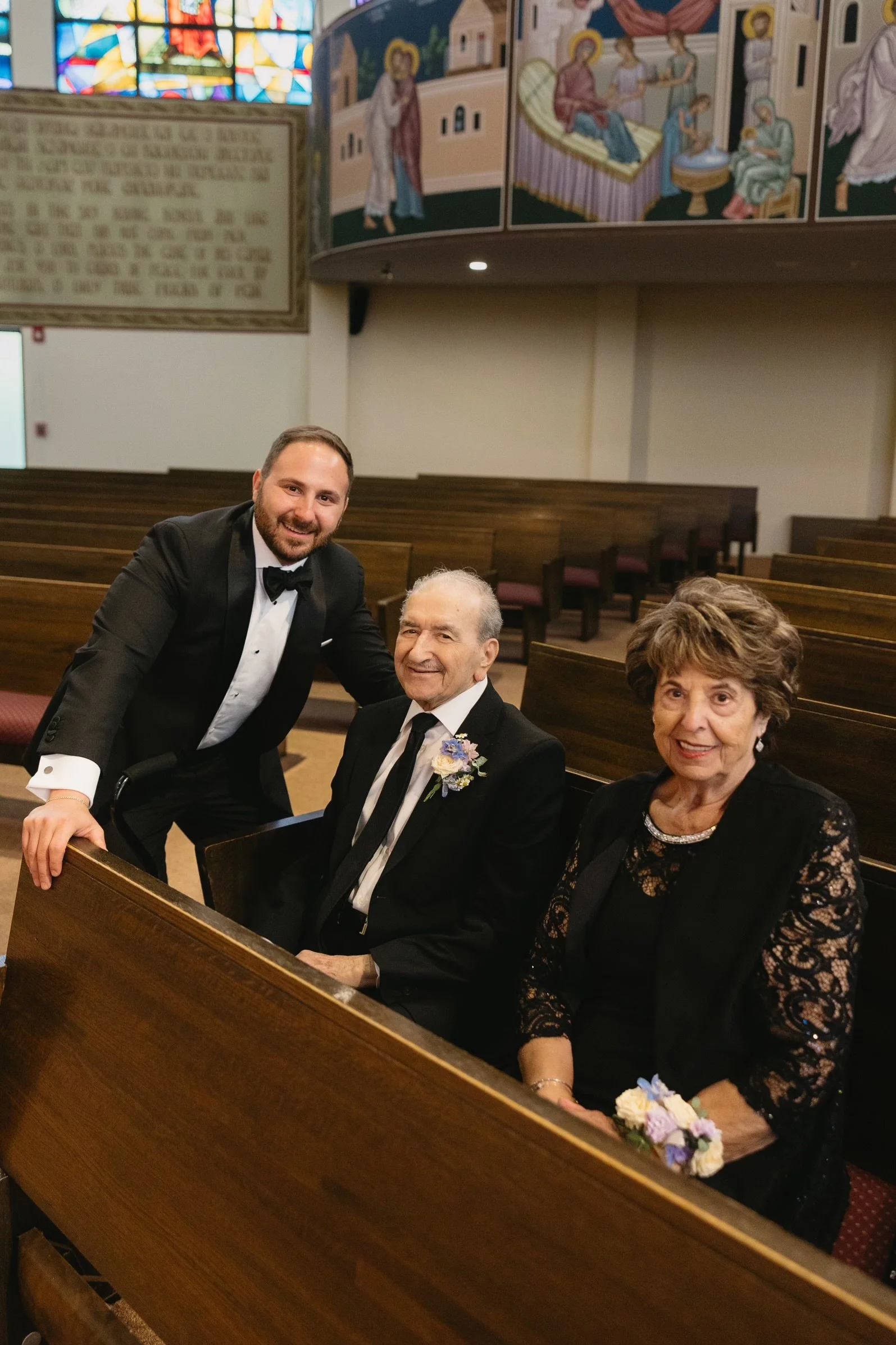 A young man in formal tuxedo poses with an elderly man and woman inside a church, with the elderly man sitting on a pew and the woman sitting beside him, all smiling for the camera.