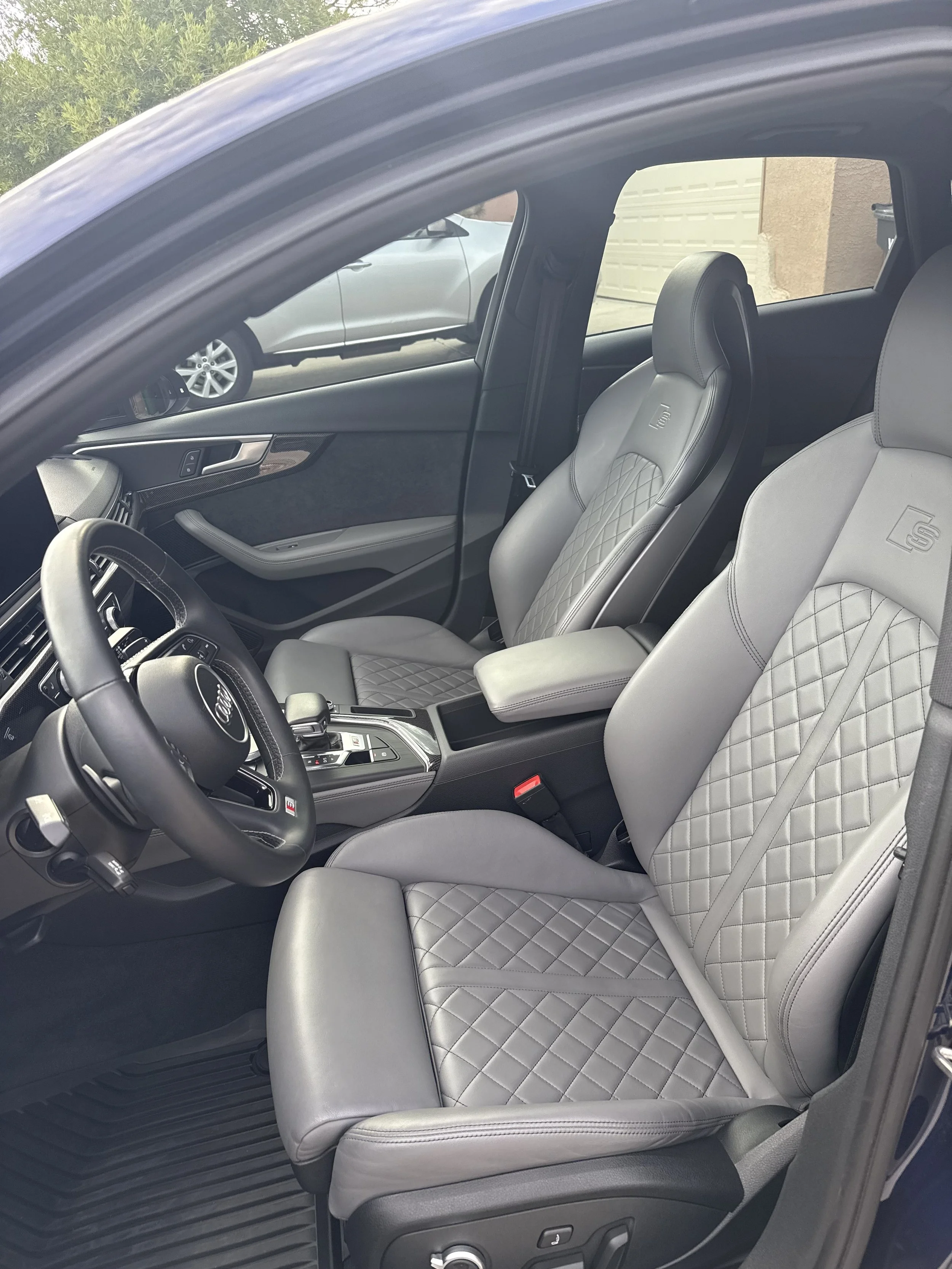 Interior of a car showing gray leather seats with diamond stitching, a black steering wheel, and control buttons on the center console. The dashboard and door panel are also visible.