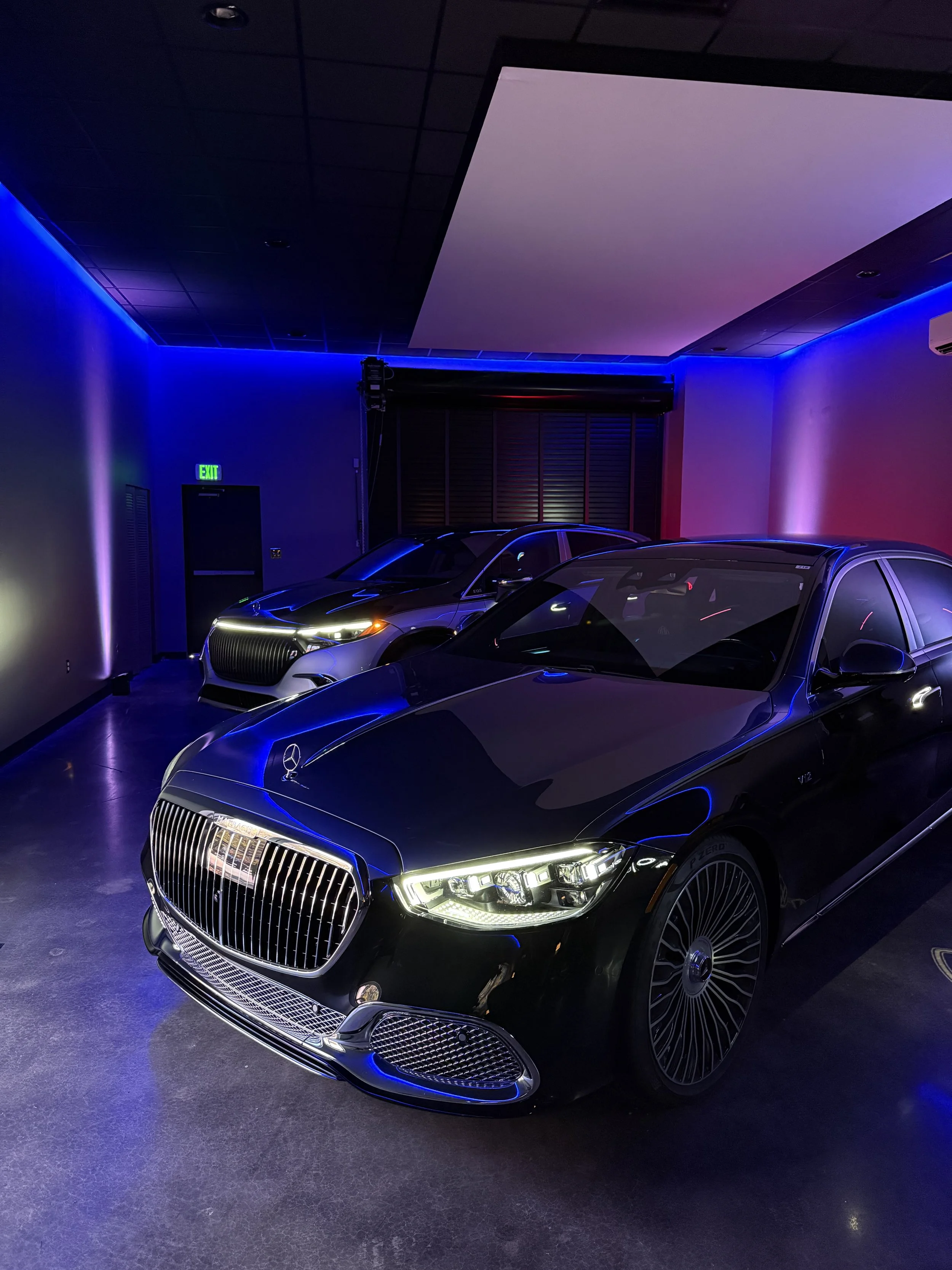 Black luxury car with a large chrome grille and LED headlights, parked indoors with purple and blue ambient lighting.