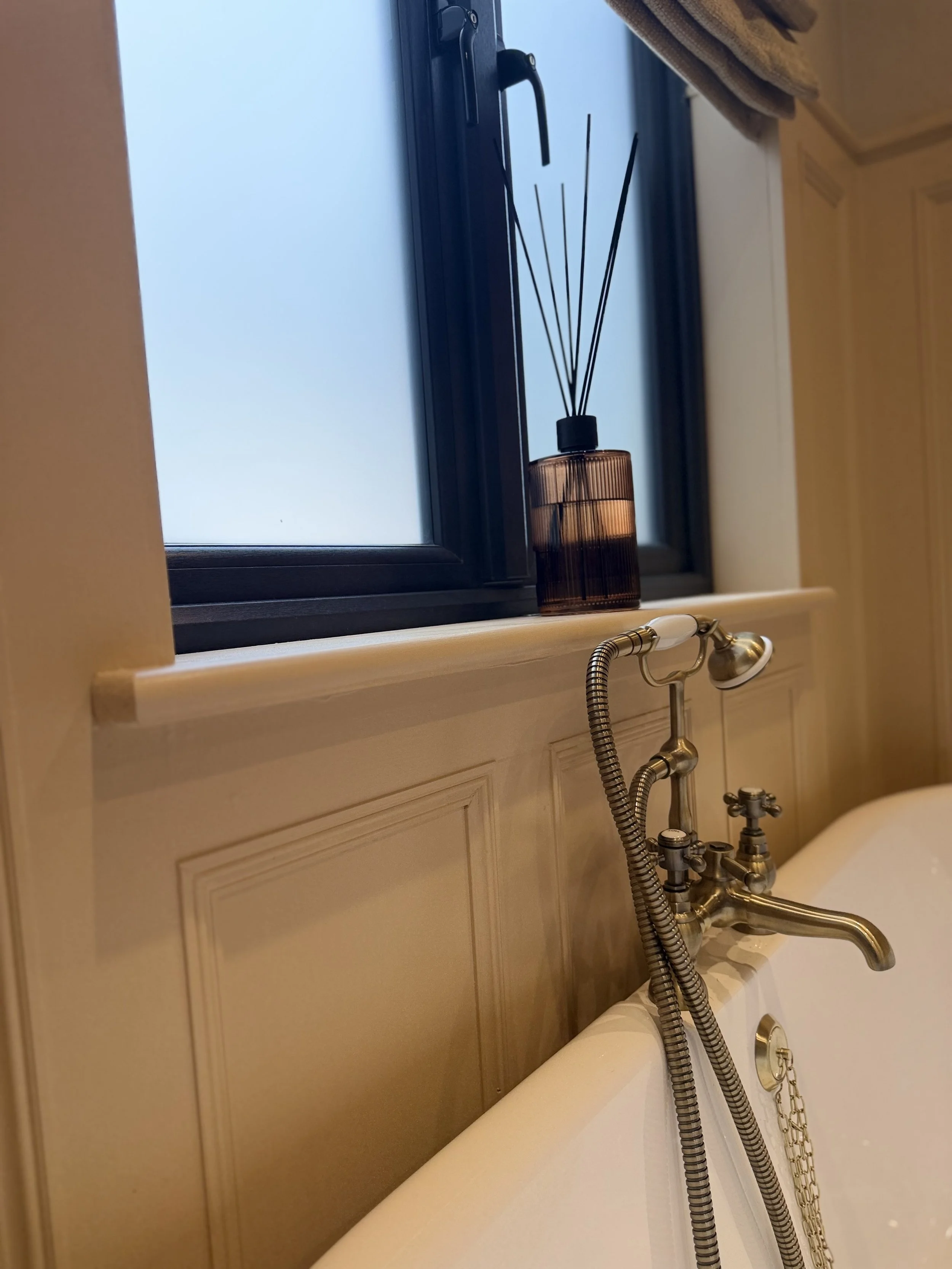 Close-up view of a bathroom window with a brown scent diffuser, a shower head, and a faucet on a white bathtub with beige cabinetry.