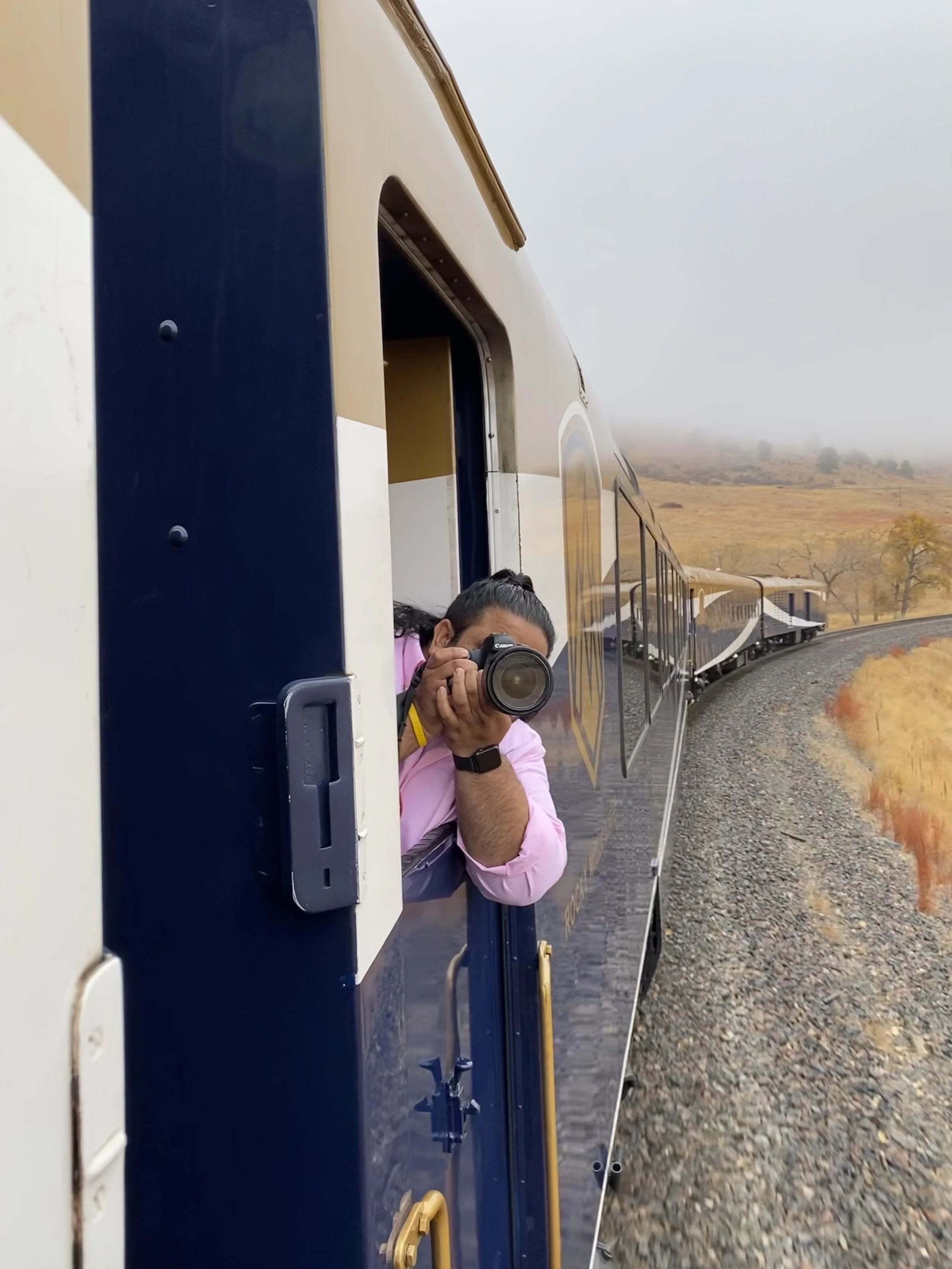 A person with a camera taking a picture from a train window with a landscape view of trees and hills in the background.