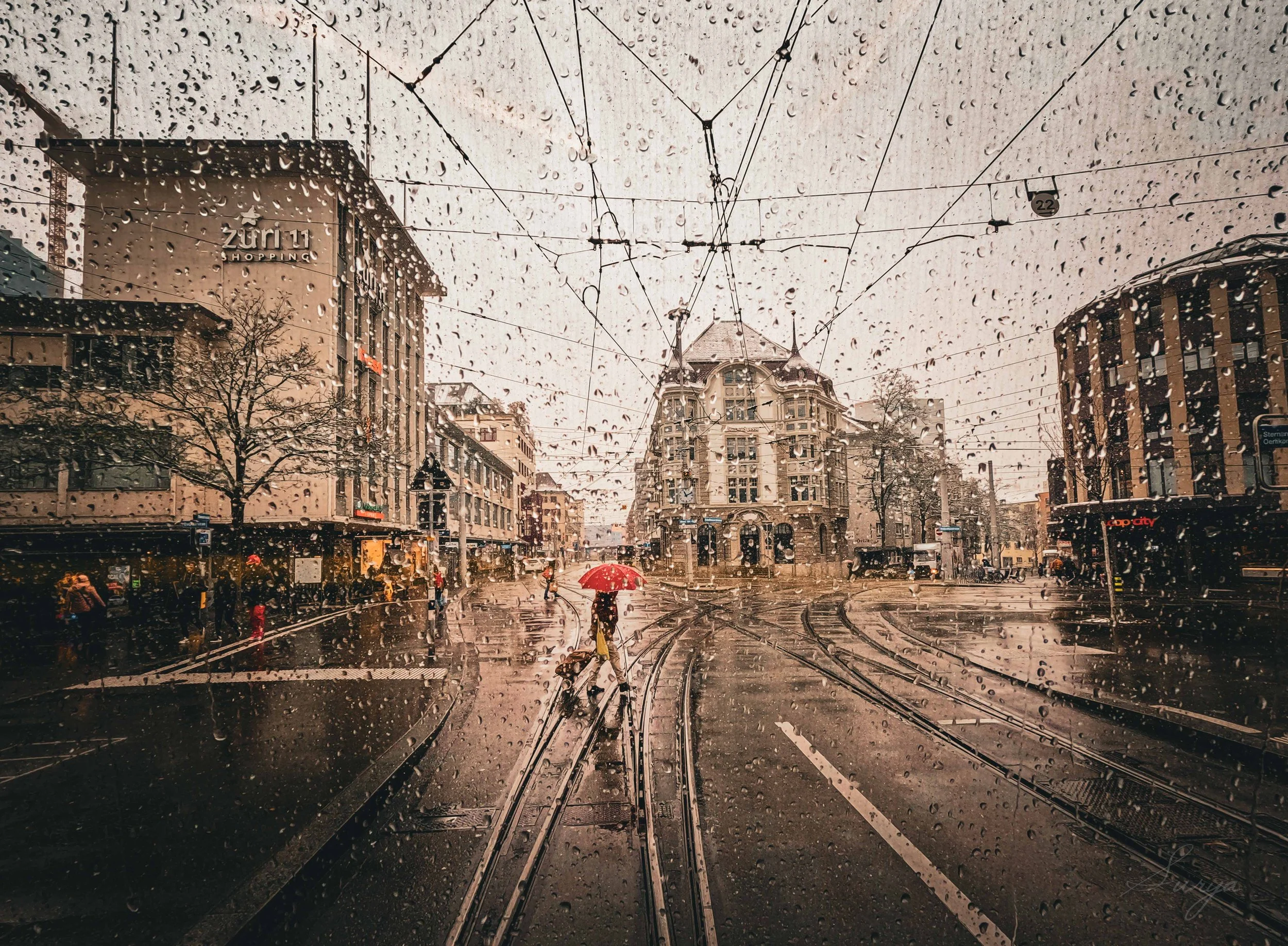 View through a rain-covered window of a city street with tram tracks, buildings, trees, and pedestrians holding umbrellas, on a rainy day.