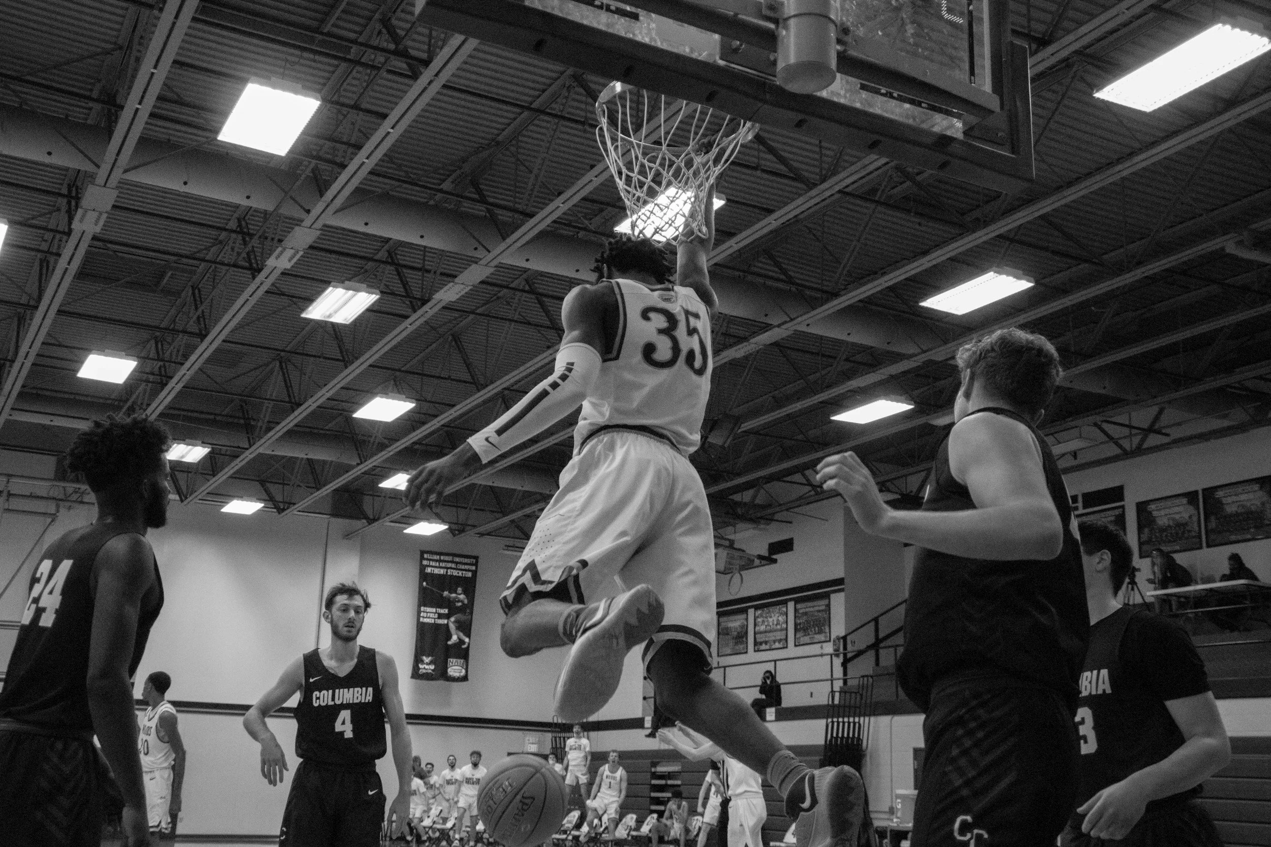 A basketball game is taking place in an indoor gym. A player wearing jersey number 35 is in mid-air performing a slam dunk, with the ball just about to go through the hoop. Other players are watching or preparing for the play. The ceiling has bright ceiling lights, and there are banners and framed pictures on the walls.