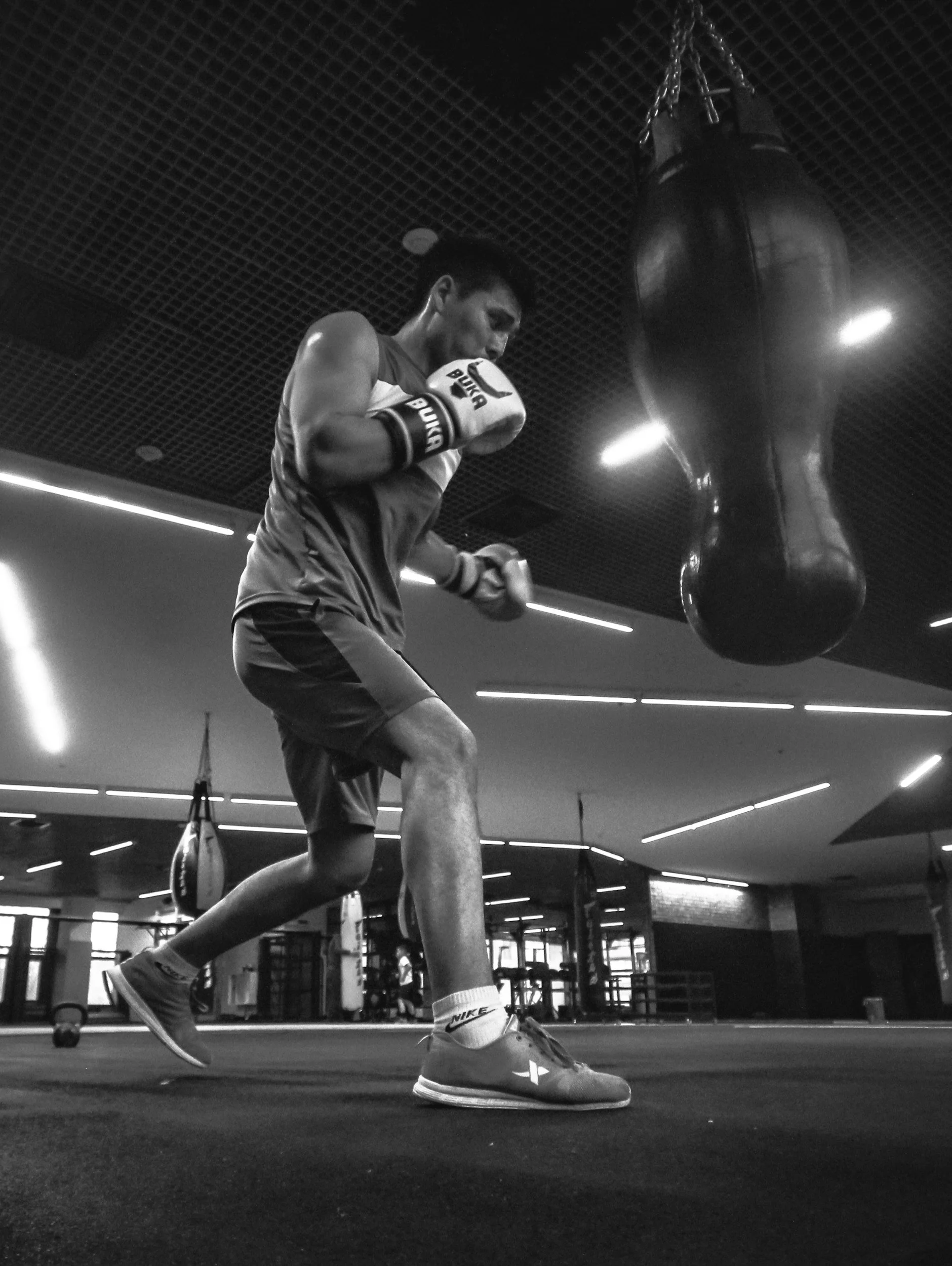 A boxer in athletic gear practicing on a punching bag in a gym, with the gym's modern ceiling lights visible.