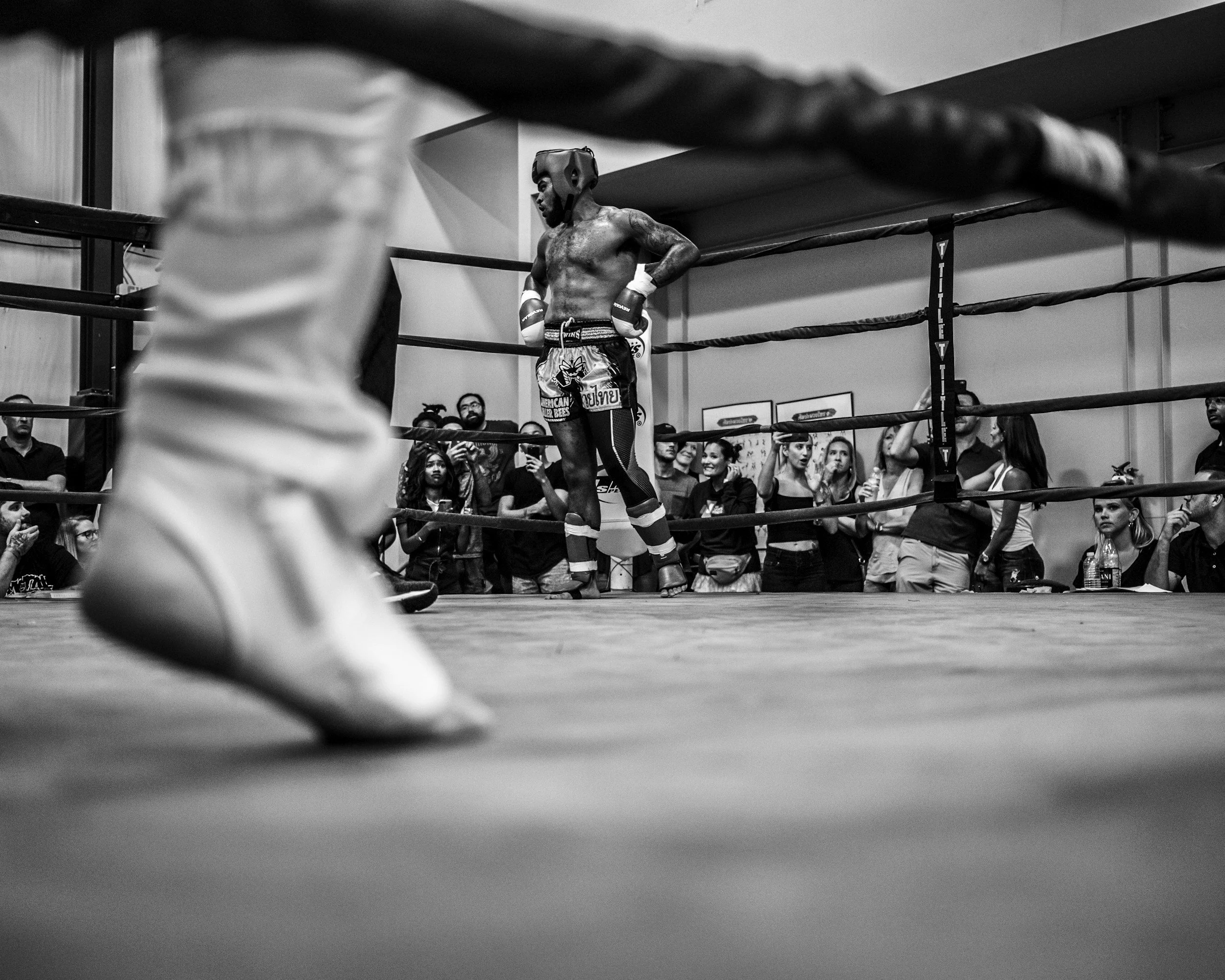 Black and white photo of a boxer standing in a boxing ring, oxygen helmet on his head, surrounded by spectators in the background, with the focus on the boxer's muscular torso and gloves.