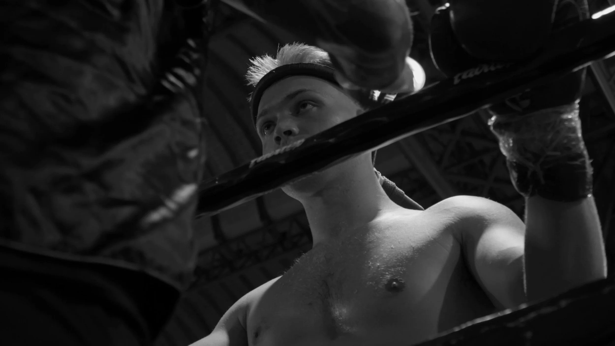 A black and white photo of a boxer in the ring, looking up through the ropes with a serious expression, wearing gloves and a headband.