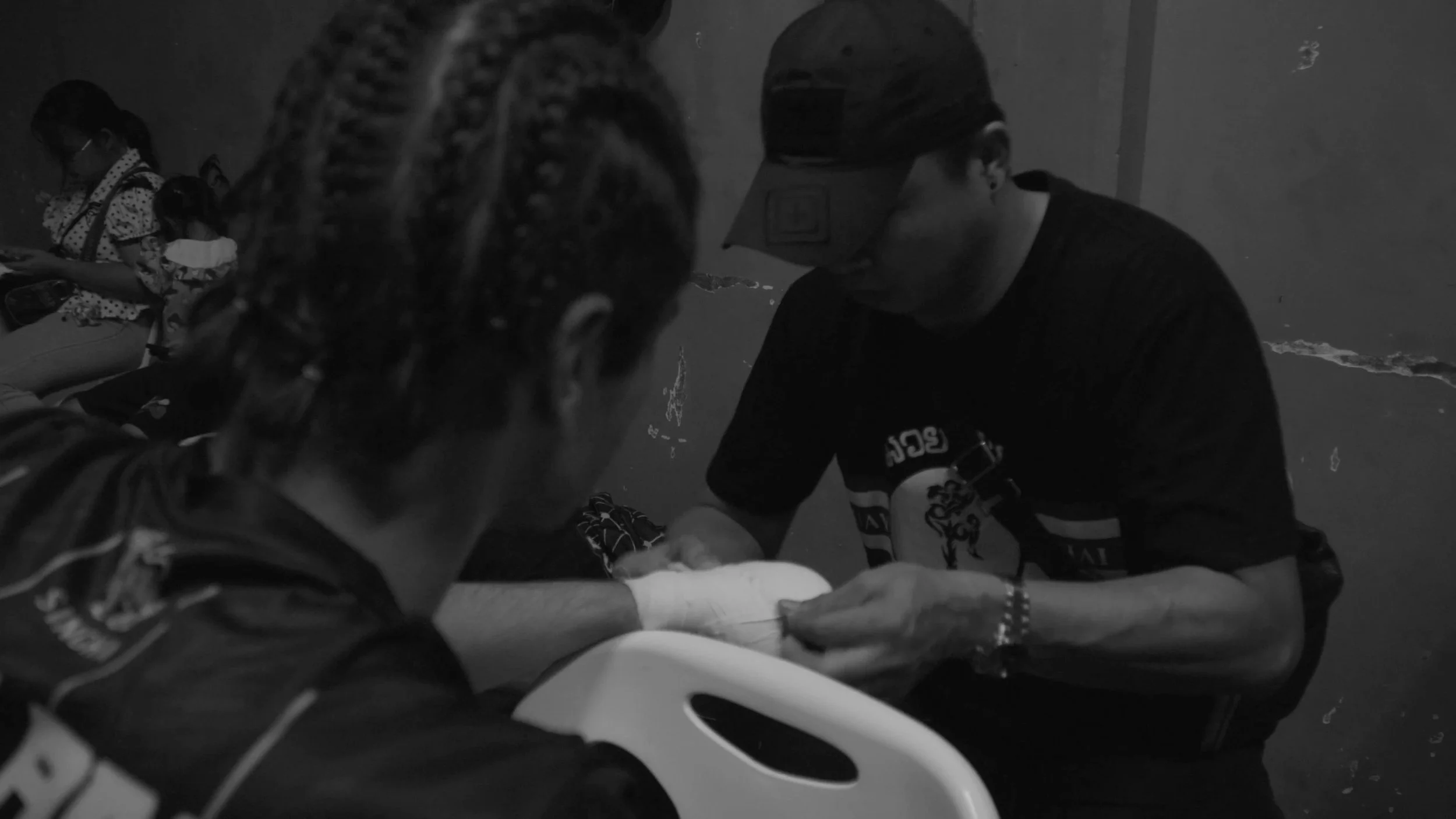 A black and white photo of a man wearing a cap and glasses treating a person's hand, with a woman sitting in the background looking at her phone.
