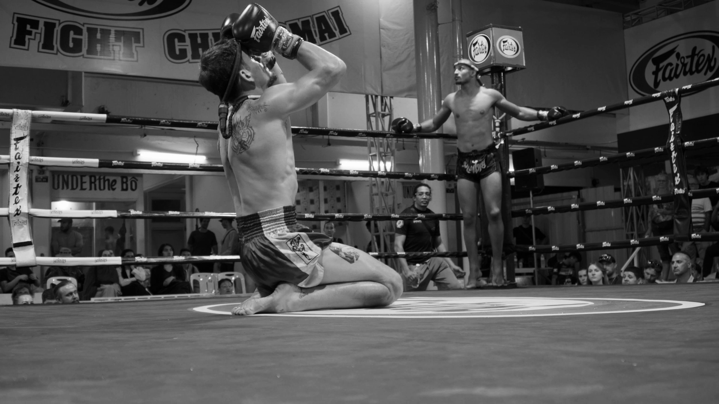 A shirtless martial artist kneeling in a boxing ring, with his hands raised to his forehead. In the background, a referee and a boxer on the ropes observe, with an audience watching the scene.