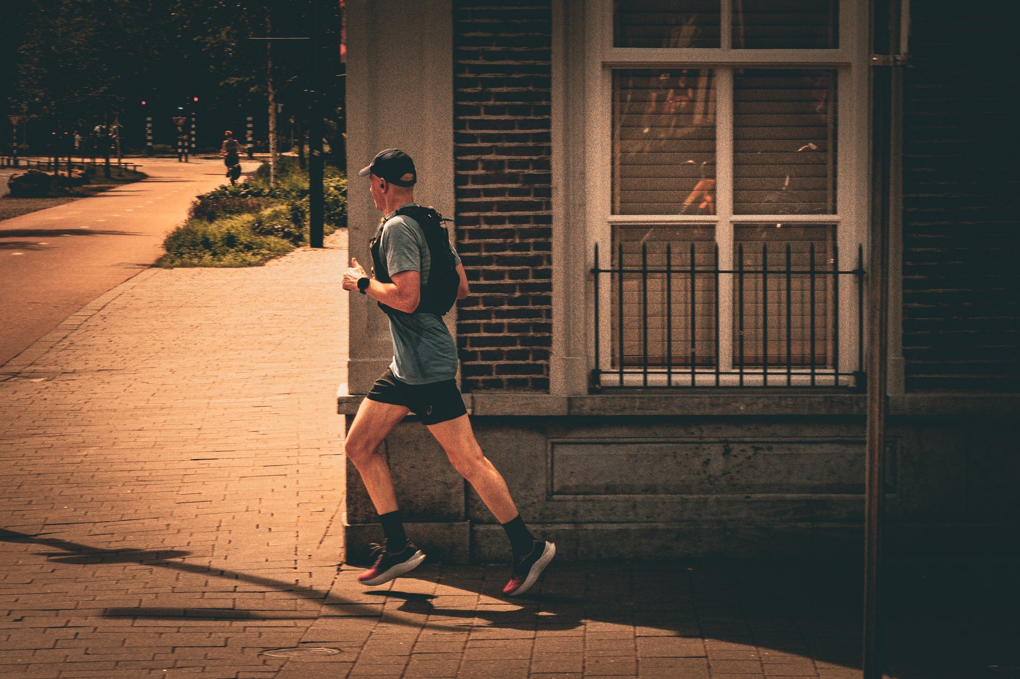 Een man in sportkleding rent 's avonds langs een huis met een grote raampartij en een balkon, onder een donkere hemel.