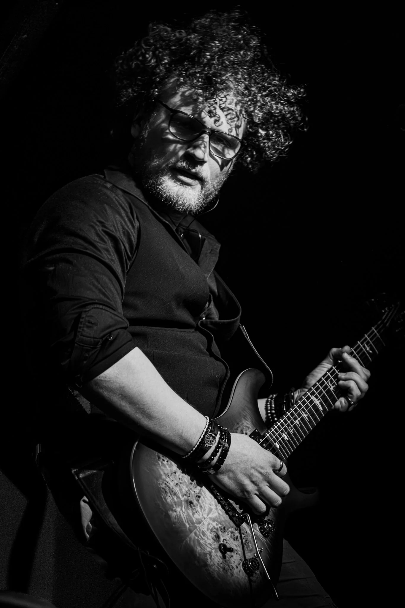 Man met een gitaar, opgespannen in zwart-wit, met krullend haar en een bril, die lijkt te zingen of praten. Donkere achtergrond.