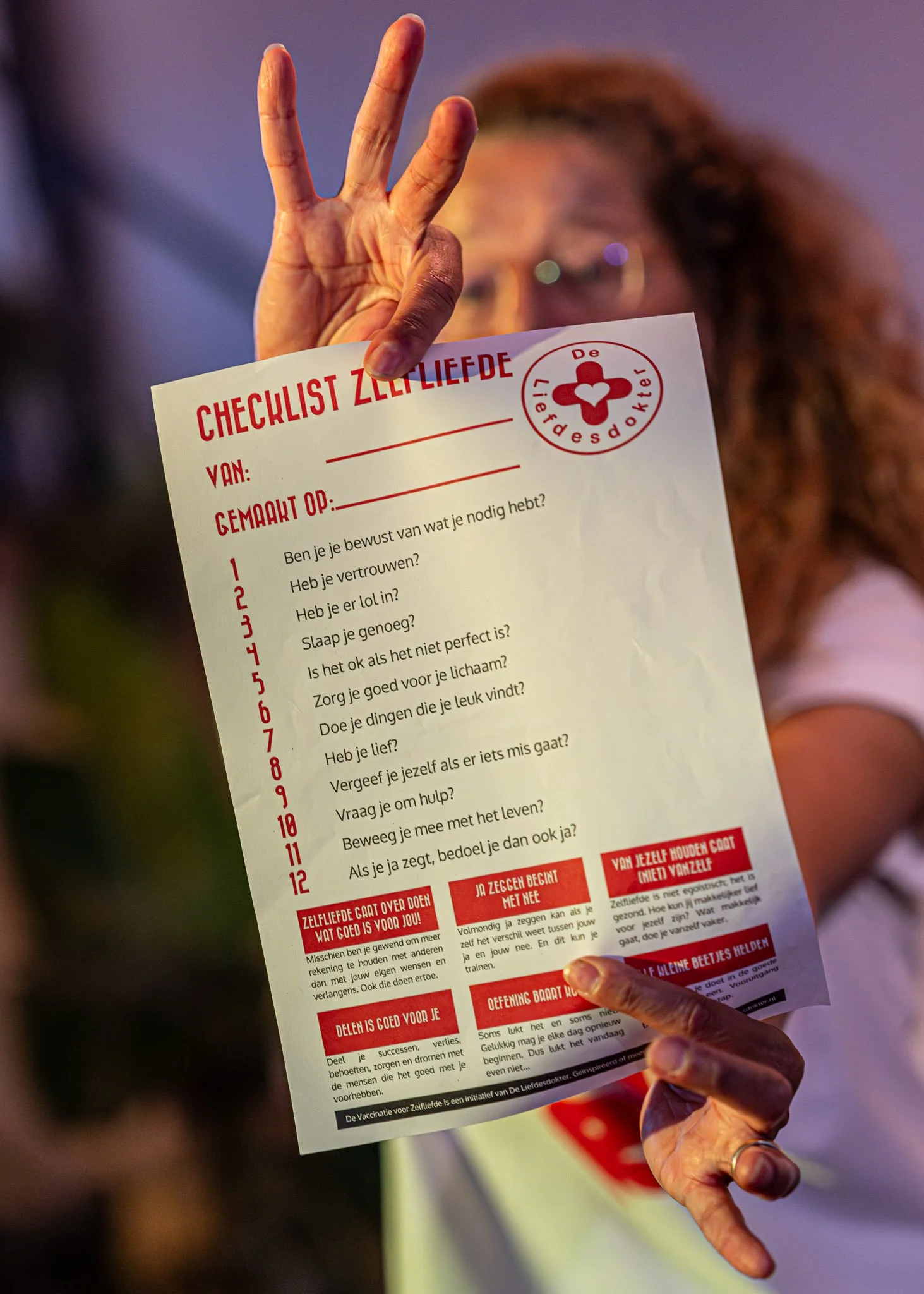 Een hand houdt een Nederlandse flyer met de titel 'Checklist Zelfliefde' en een lijst met vragen, onder een logo dat zegt 'De Liefdesdokter', op de achtergrond zijn andere mensen die mogelijk deelnemen aan een workshop of evenement.