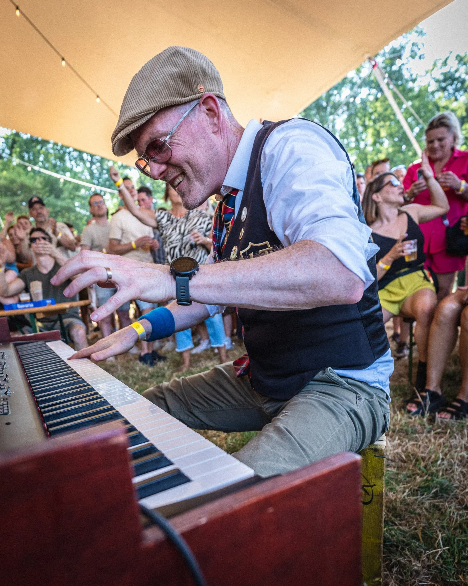 Een man speelt keyboard tijdens een buitenmuziekfestival onder een grote oranje tent met een groep mensen op de achtergrond.