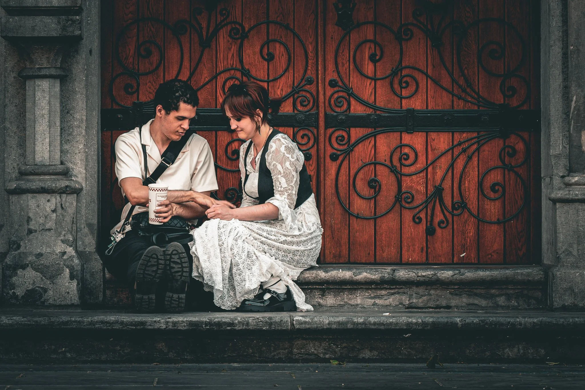 Twee mensen zitten op een traptrede voor een houten poort met ijzeren sierroedes. Het is een man en een vrouw die samen naar elkaar kijken. De vrouw draagt een witte kanten jurk en de man draagt een lichte blouse. Ze lijken dicht bij elkaar te zitten
