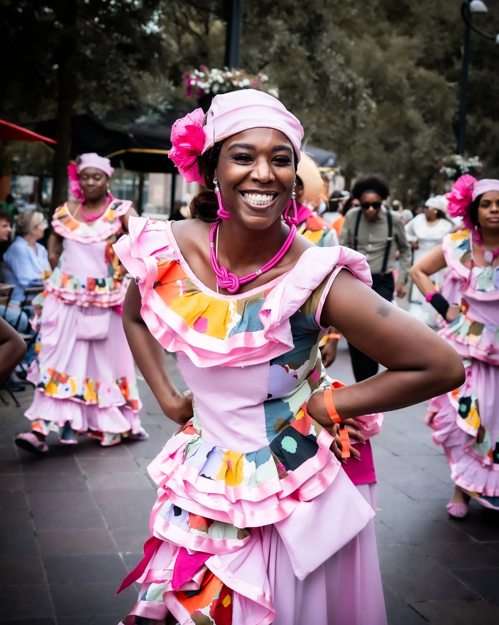Vrouw in kleurrijke traditionele danskostuum danst tijdens een feest of parade op straat. Ze draagt een roze outfit met bloemenmotief, grote roze oorbellen, en een roze hoofdband met bloemen.