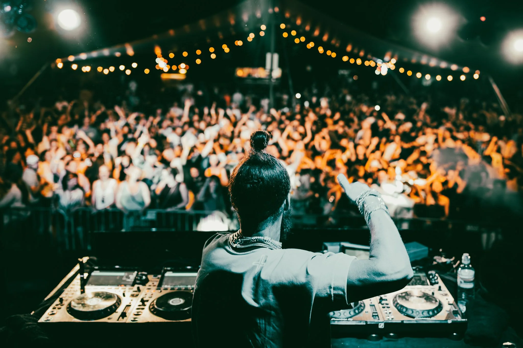 DJ die op een draaitafel in een tent met een groot danserpubliek zich bevindt, kleurrijke lampjes hangen boven de menigte.