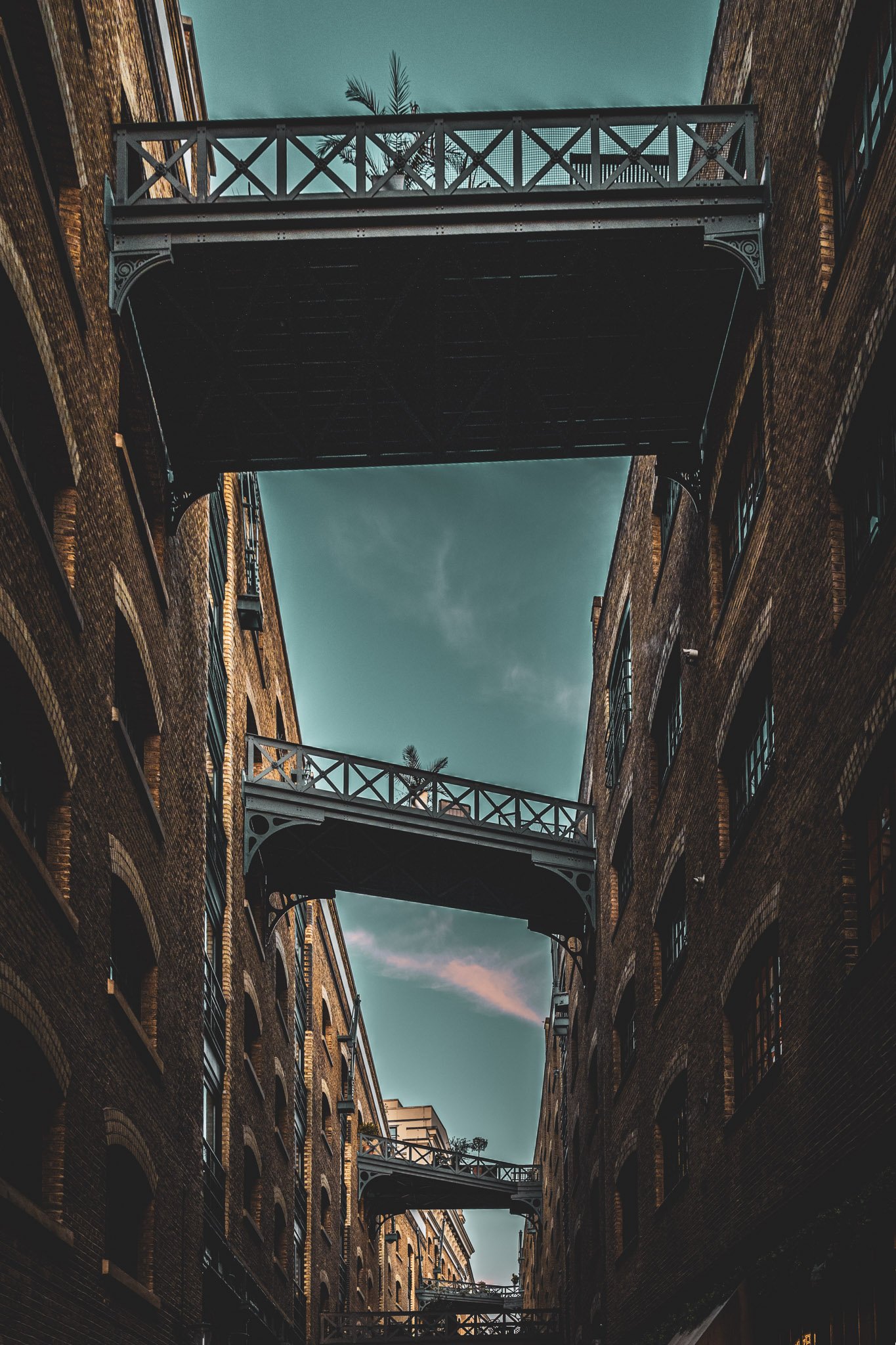 Fotografie van een smal straatje tussen oude bakstenen gebouwen met metalen balkons en een hemel met wolken op de achtergrond.