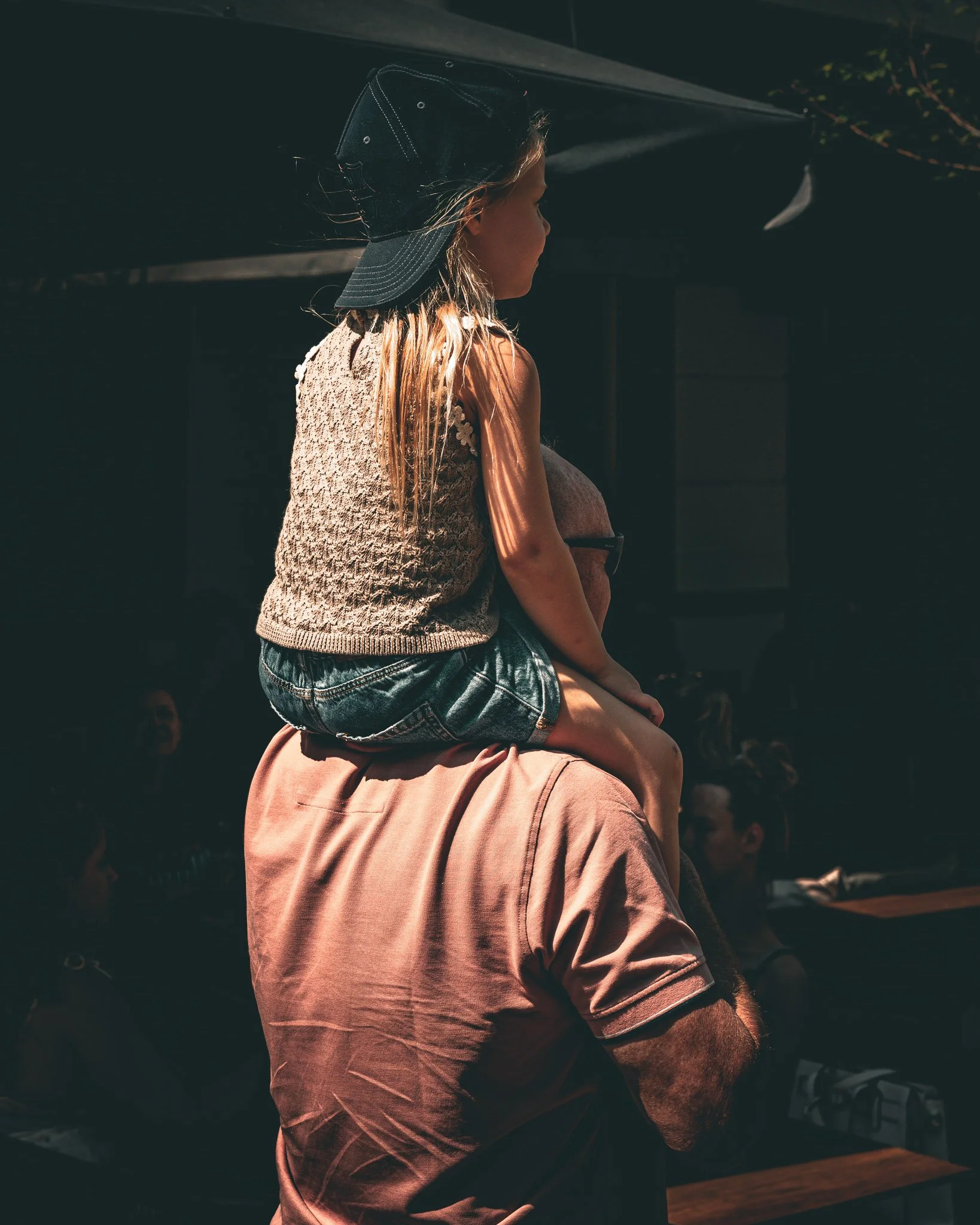 Een jonge vrouw met lang blond haar zit met een jongen op de schouders in een drukke omgeving, mogelijk tijdens een evenement of festival. Ze dragen zomerkleding, en de situatie lijkt in de buitenlucht.