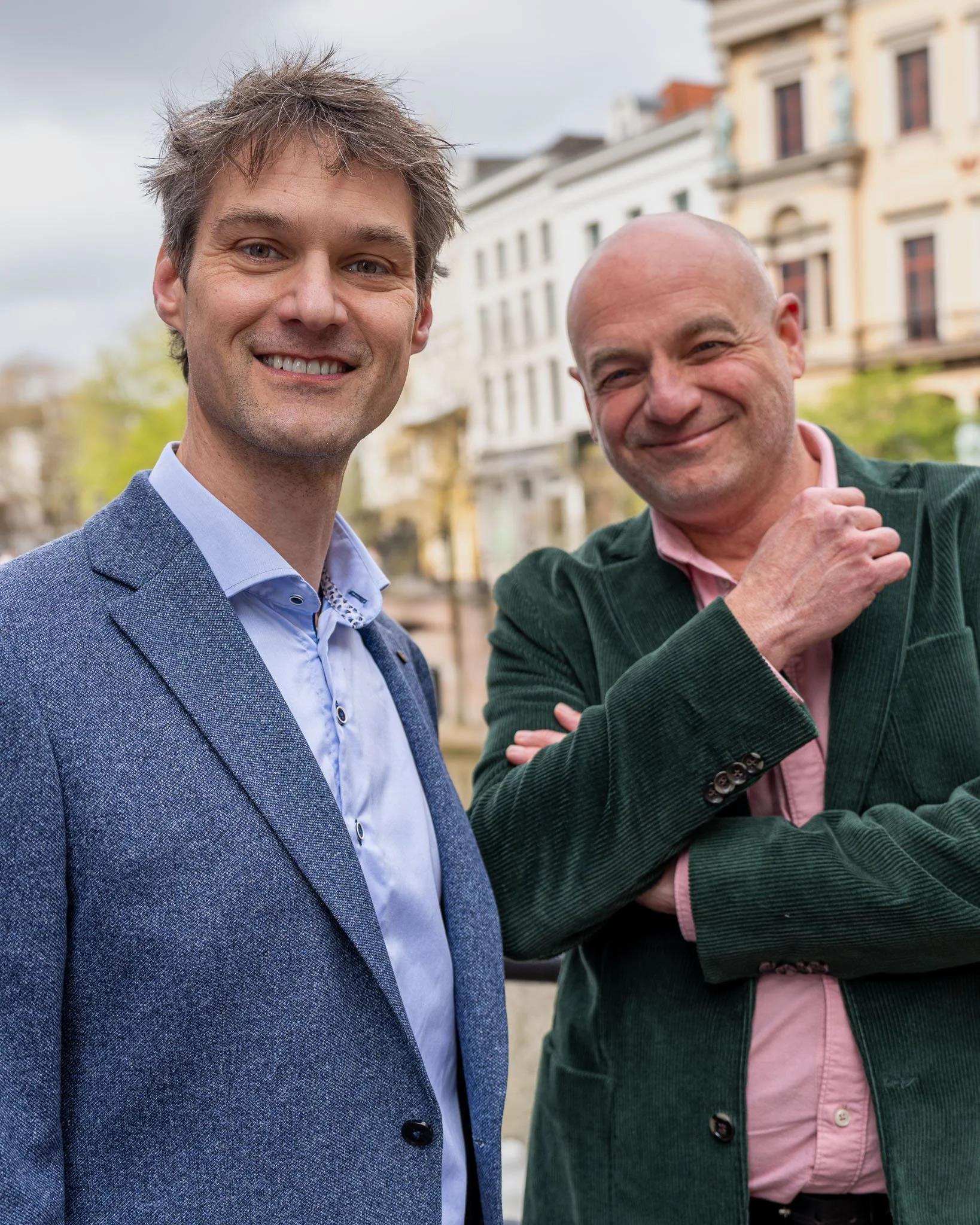 Twee lachende mannen in een stad met historische gebouwen, een man met bruin haar en een blauwe blazer, de andere met een kale kop en een groene jas.