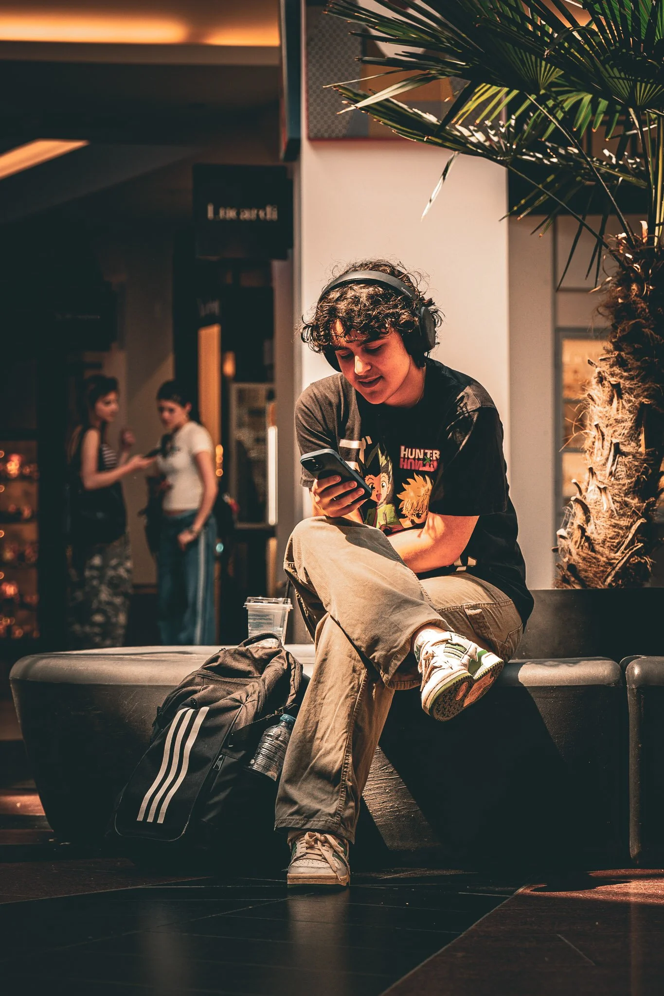 een jonge man met curly haar zit op een bankje in een binnenruimte, draagt een koptelefoon en is bezig met zijn mobiel, er ligt een tas en een waterfles voor hem, op de achtergrond twee vrouwen die praten, een palmboom en warme verlichting.
