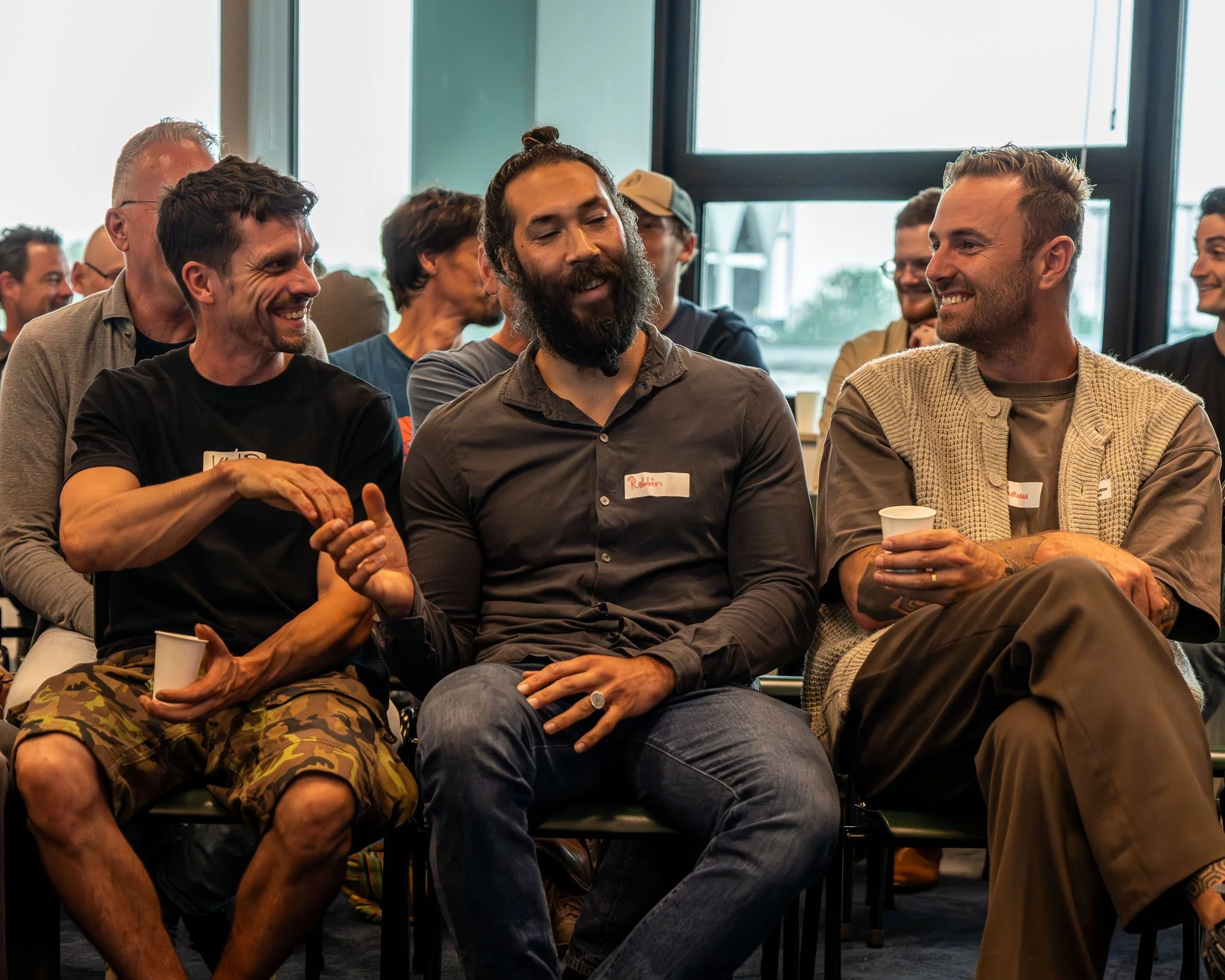 Vijf mannen en vrouwen die lachen en praten, zittend op stoelen tijdens een evenement in een kamer met grote ramen.