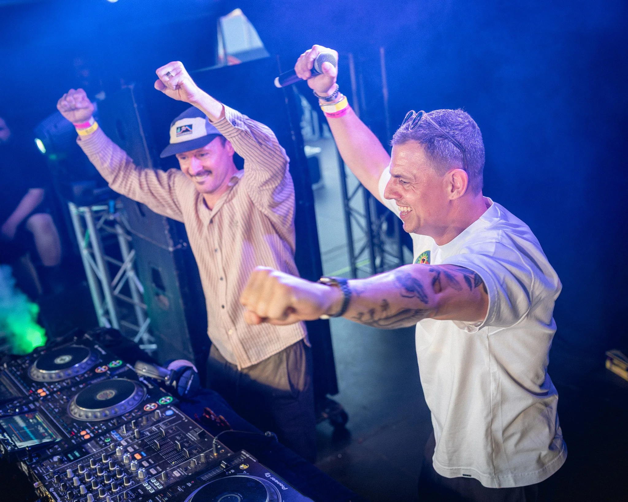 Twee mannen op een dansfeest, een draaitafel en DJ-apparatuur op de voorgrond, één man met een hoofddeksel die danst en de ander met een horloge die de handen omhoog steekt.