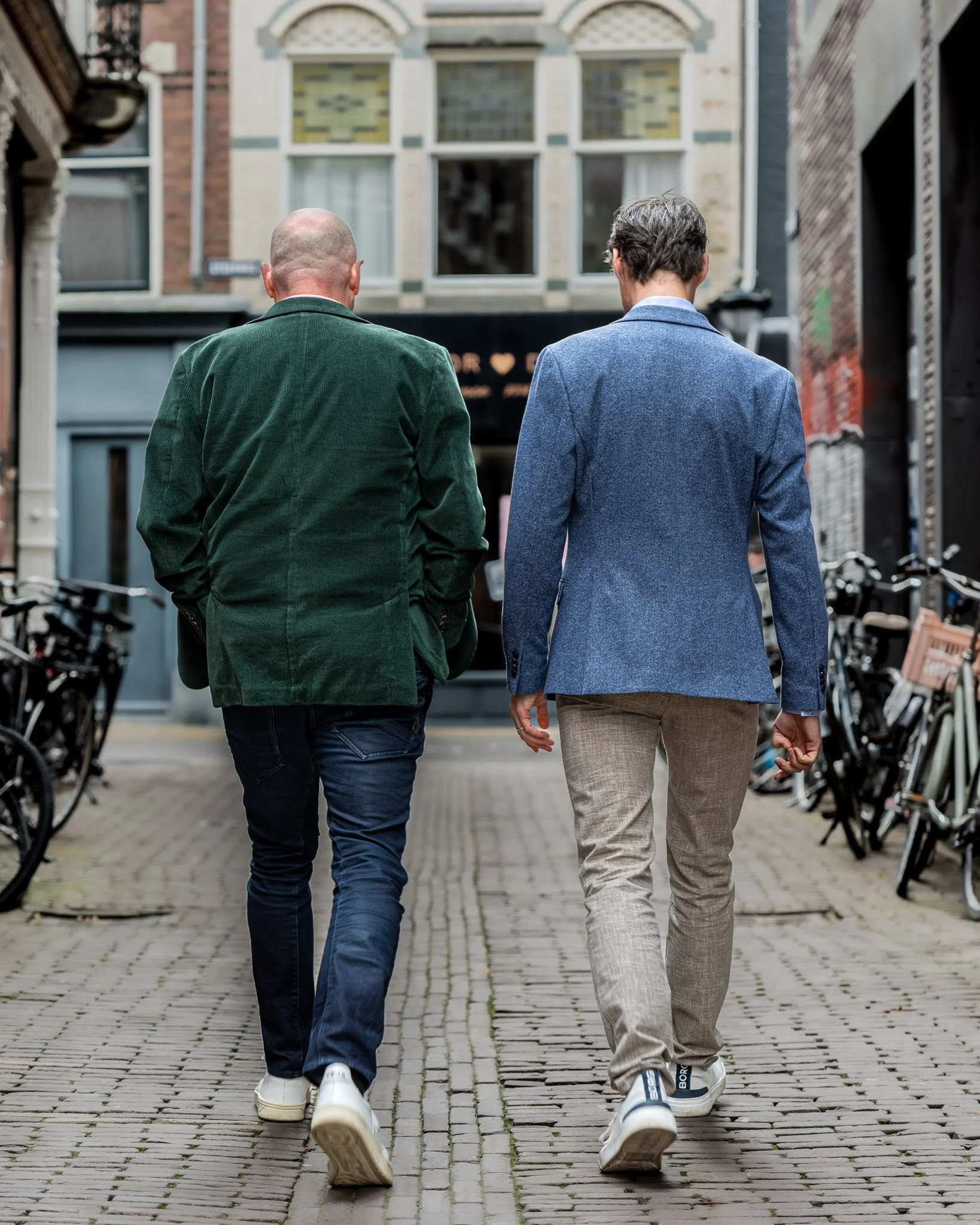 Twee mannen lopen samen op een stenen straat in een stedelijke omgeving, met fietsrekken en gebouwen aan weerszijden.