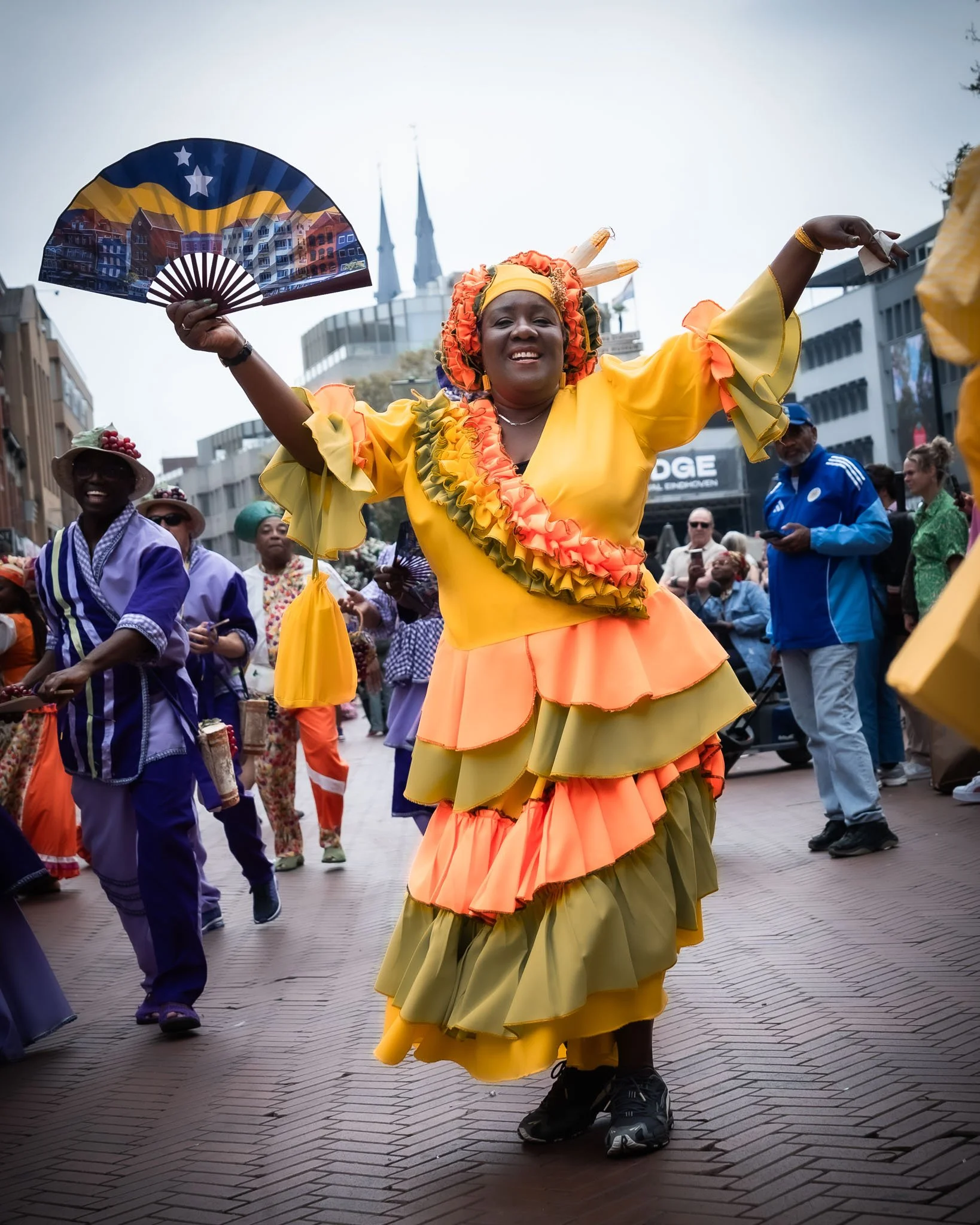 Vrouw in kleurrijk folklore kostuum danst tijdens parade op straat, omringd door publiek en andere deelnemers.