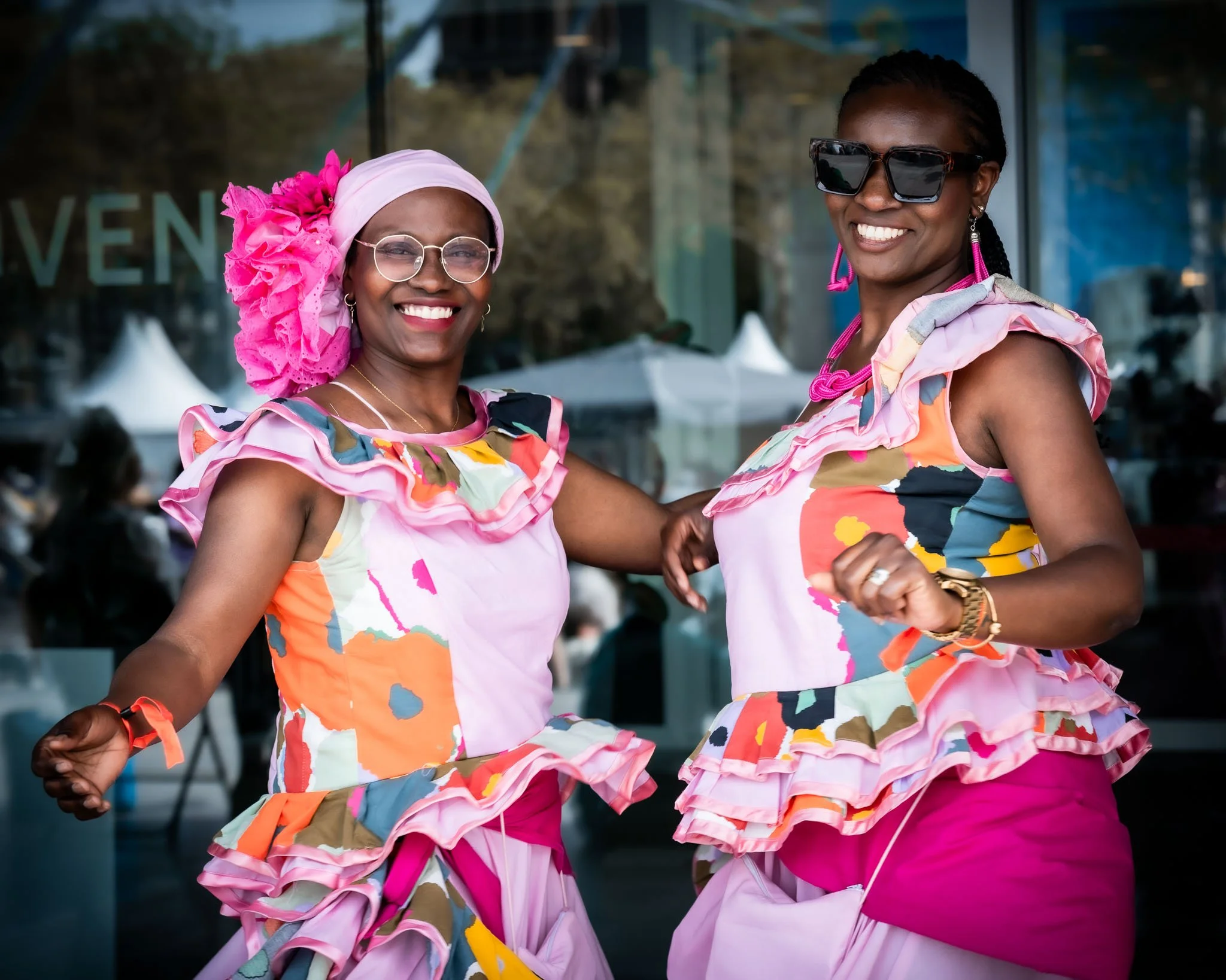 Twee vrouwen dansen samen, gekleed in kleurrijke kleding met patronen, in een buitenomgeving met een glazen gebouw op de achtergrond. Ze lachen en dragen accessoires zoals zonnebrillen en oorbellen.