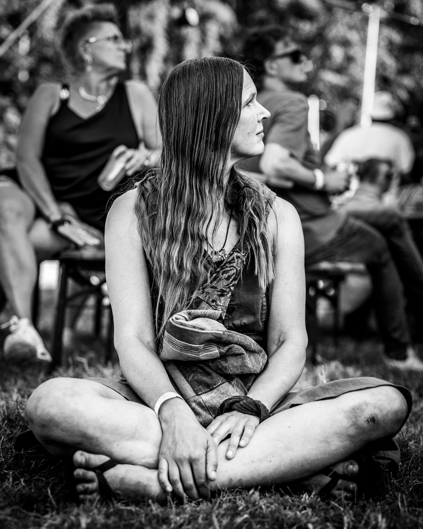 Een vrouw zit in een kring met andere mensen, haar benen gekruist en haar armen over haar knieën, in een natuurlijke omgeving. Ze kijkt aandachtig naar rechts.
